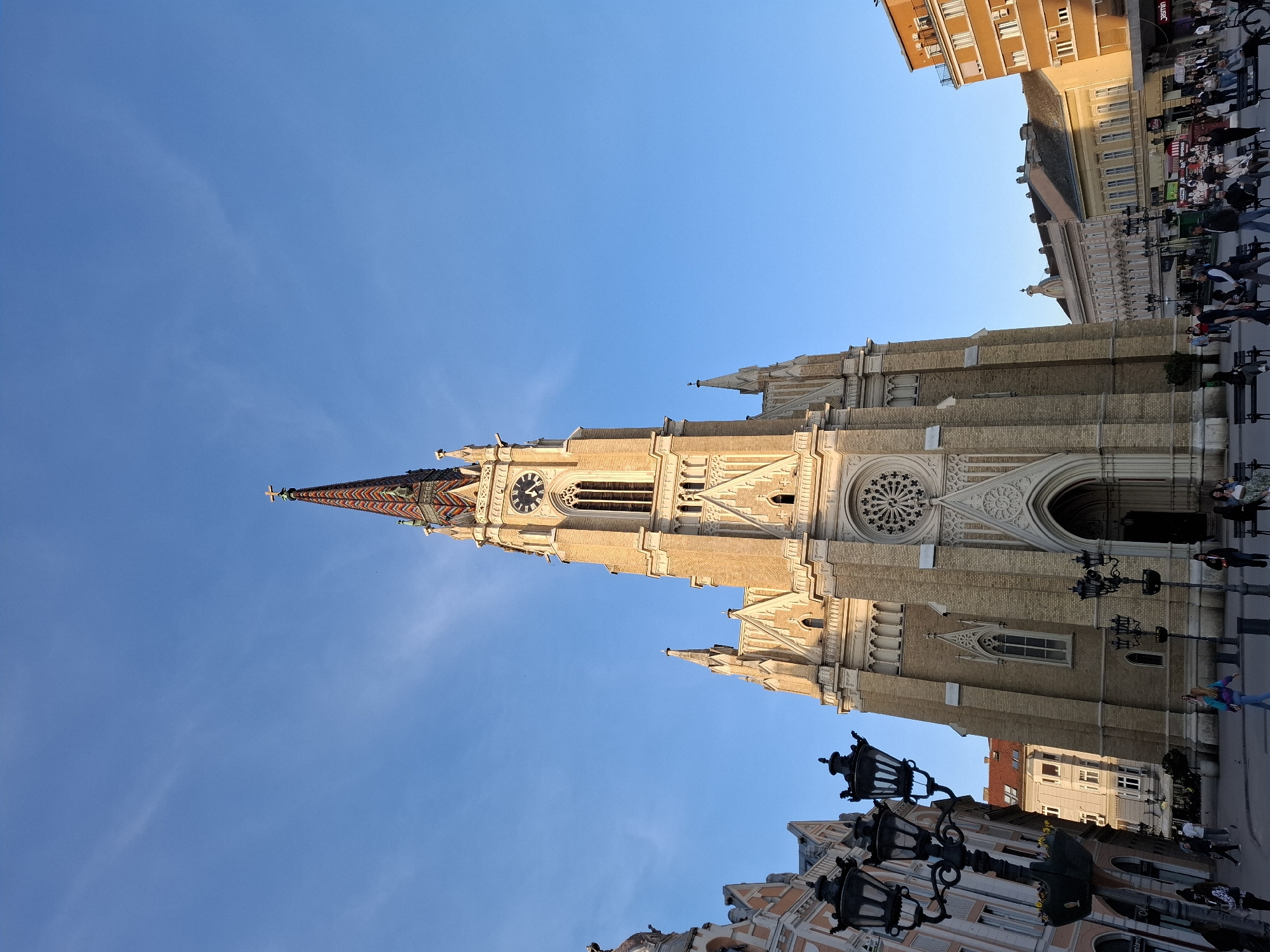 The image depicts the Name of Mary Church, a Roman Catholic parish church in Novi Sad, Serbia, from a low angle, emphasizing its height against a clear blue sky. The church features a tall spire with a colorful patterned roof, a clock tower, and a rose window. The church is constructed of light-colored brick. People are visible on the street beside the church. Buildings of various architectural styles are visible. Street lamps are placed along the street.