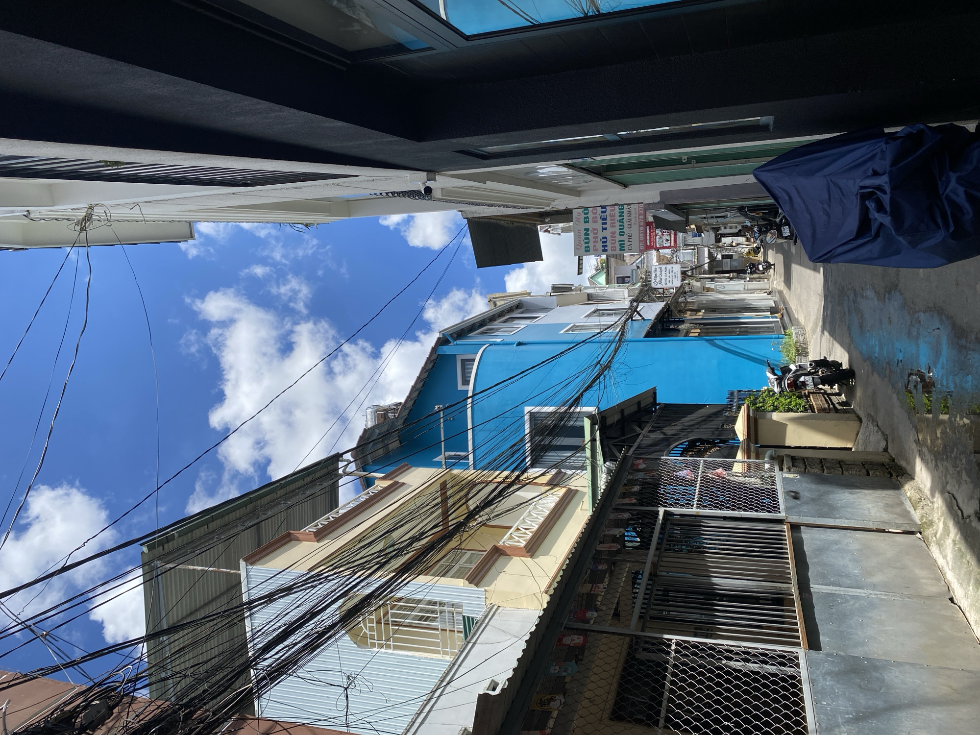 This is a low-angle, full-shot photograph taken on a sunny day in a narrow alleyway in the city of Cam Ly, Đà Lạt, Vietnam. The alley is flanked by buildings of various colors, including a striking bright blue building on the left and a predominantly black building on the right. The sky is a vibrant blue, with scattered white clouds. A dense network of electrical wires crisscrosses overhead, creating a complex visual texture against the sky.
On the left side of the alley, a light yellow building with brown trim features a window with a decorative white metal grill. Below this building, a chain-link fence encloses a courtyard with several metal doors. Hanging decorations, possibly small figures, are visible along the top of the fence.
The alley floor is paved with concrete, and there are visible puddles of water, indicating recent rain or a spill. A motorcycle, partially covered by a dark blue tarp, is parked on the right side of the alley. Further down the alley, more motorcycles are parked, and a few individuals can be seen walking or riding them in the distance. Signage is visible on the buildings, suggesting commercial establishments, with Vietnamese text indicating food and drink services. The overall impression is of a densely populated urban environment with a mix of residential and commercial structures, characterized by overhead wires and practical, everyday elements.