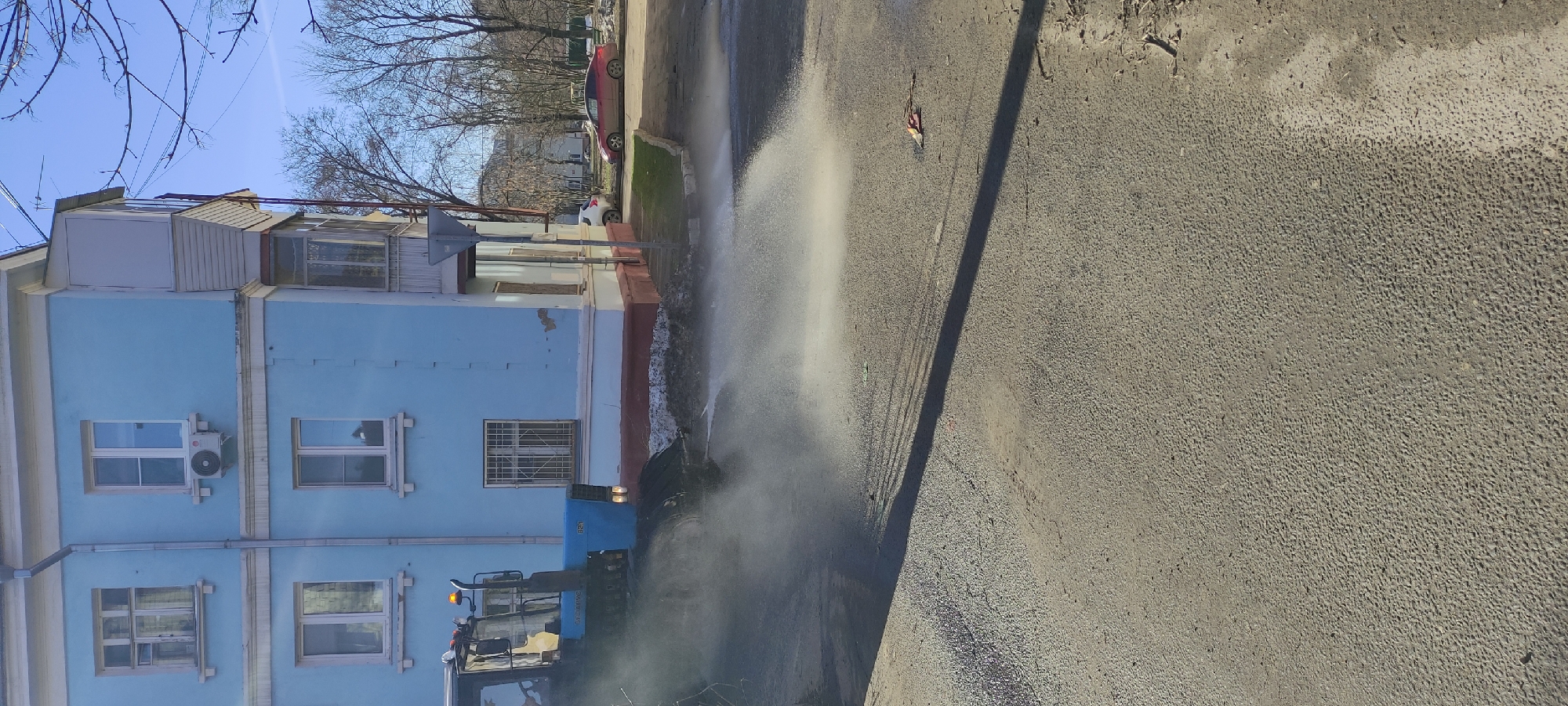 The image shows a street in Fryazino, Russia being cleaned by a blue tractor with a water spraying attachment. The tractor is positioned on the left side of the frame, next to a light blue, multi-story building with several windows. The water being sprayed by the tractor creates a visible mist or spray on the road. The road is paved and stretches across the right side of the frame. The sky is clear and blue. Trees with bare branches are visible in the background. Several parked cars are near a building on the left.
