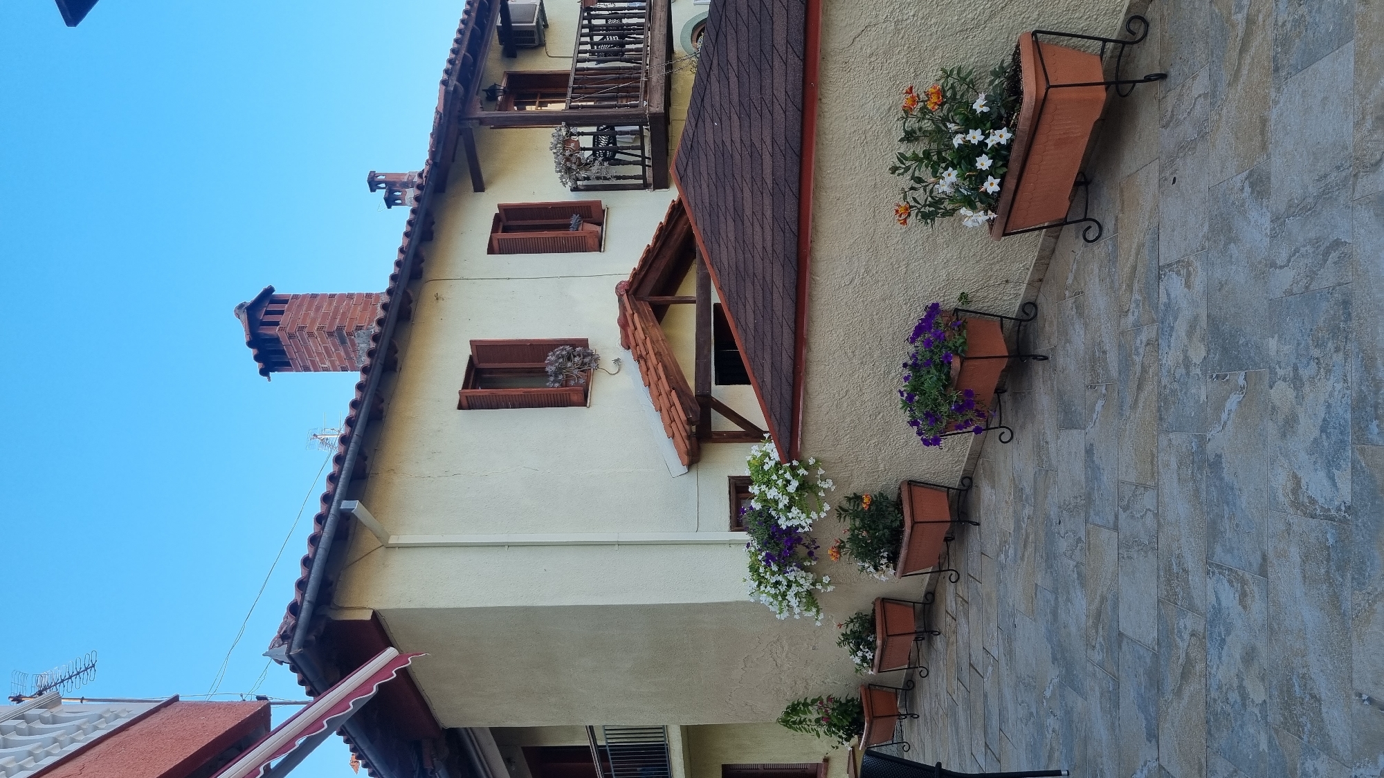 The image captures a sunlit facade of a building in Litochoro, Greece. The building, painted a light cream color, features a red-tiled roof and several windows with wooden shutters, some of which are open. Two chimneys made of red brick stand on the rooftop, indicating traditional architecture.
In front of the building, a stone-tiled patio is lined with several terracotta planters filled with vibrant flowers. White, purple, and orange blooms create a colorful display against the textured cream wall. A balcony with a wooden railing extends from the upper floor, where outdoor chairs are visible. The clear blue sky suggests a pleasant weather day. The overall scene evokes a serene and picturesque atmosphere typical of a Greek coastal town.