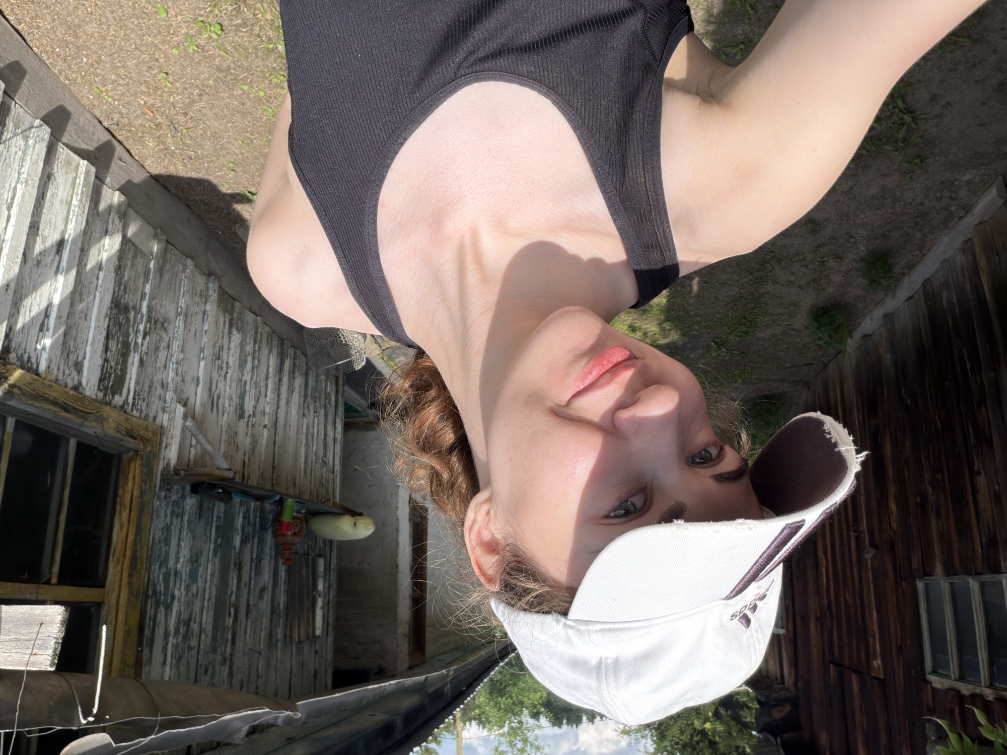 In Zavodoukovsk, Russia, a young woman with light skin and curly brown hair tied back wears a white baseball cap and a black tank top. She is outdoors, posing for a selfie in front of a weathered wooden building with a corrugated metal roof. The building appears rustic, with peeling paint and a slightly tilted, makeshift awning above a window. In the background, a wooden fence lines the yard. The ground is covered in dry dirt and sparse green vegetation. On a shelf attached to the building, a few objects are visible, including what appears to be a light-colored gourd, a decorative vase, and a red spray can. The sunlight casts shadows, indicating it is daytime.