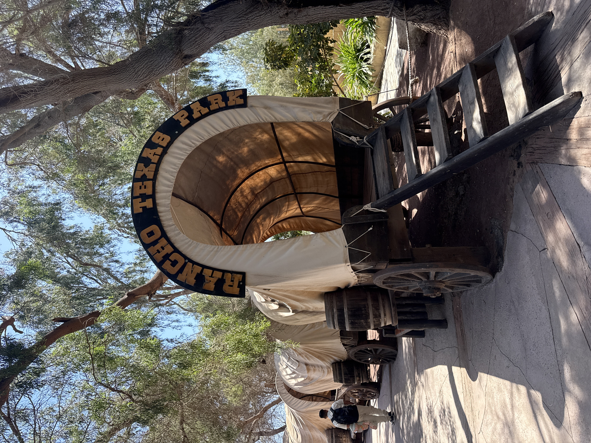 The image shows covered wagons with "Rancho Texas Park" signage. The location is likely Rancho Texas Lanzarote Park in Tías, Spain. The wagons are in an outdoor area with trees and paved ground. There is a person in the lower section of the photograph.