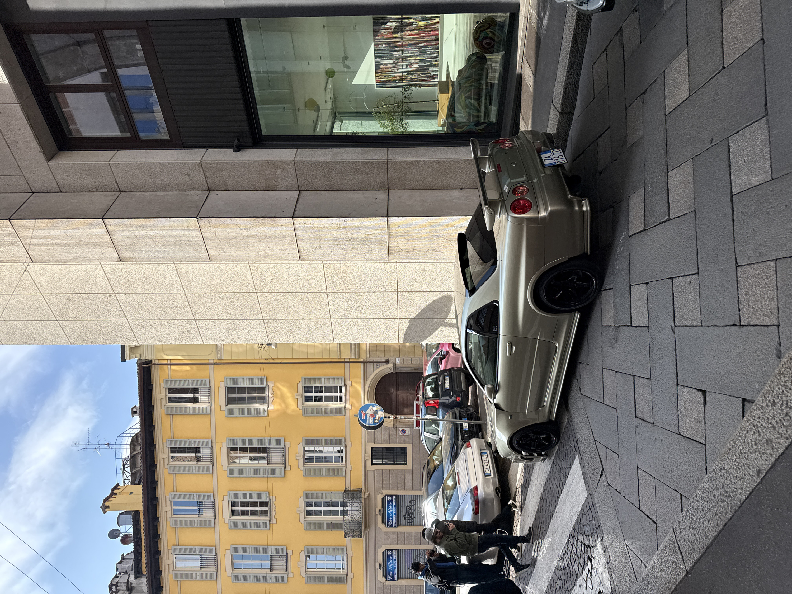 The scene depicts a street in Milan, Italy, on a clear, sunny day. A gold Nissan Skyline GT-R, identifiable by its rear spoiler, distinct taillights, black multi-spoke wheels, and license plate "ZA 015ZD," is parked on the right foreground, angled slightly toward the viewer. The vehicle rests on a street paved with dark gray rectangular stone pavers, some lighter in shade, forming an irregular pattern.
To the right of the Skyline is a modern building constructed with light-colored, textured stone panels. Its ground floor features a large, dark-framed display window reflecting portions of the opposite street and revealing interior elements: multiple suspended bird mobiles, circular decorative objects, and a patterned hippopotamus sculpture alongside a multi-colored artwork.
On the left side of the street, two adult males are walking near parked vehicles. The first male, wearing a green jacket, light baseball cap, and jeans, is observed looking at a smartphone. Behind him, another male in a dark hoodie and blue scarf holds a camera, seemingly pointed towards the parked cars.
Behind these individuals, a silver Porsche 911 (license plate "CA-414LY") is parked. Further back, a dark gray SUV and a red hatchback are also parked. A white crosswalk is partially visible on the left.
Across the street, dominating the midground and background, stands a multi-story classical yellow building with numerous windows. Each window is fitted with light gray louvered shutters, many open, and some feature small wrought-iron balconies. The ground floor displays two storefronts with blue awnings bearing the text "Bar Affari" and "CAFFETTERIA - PANINI - APERITIVI." A blue circular street sign with a red diagonal line through a white car is positioned near the yellow building. The rooftop of this building is visible against a blue sky with scattered white clouds, featuring multiple chimneys, satellite dishes, and antennas. Overhead utility lines are present across the sky.