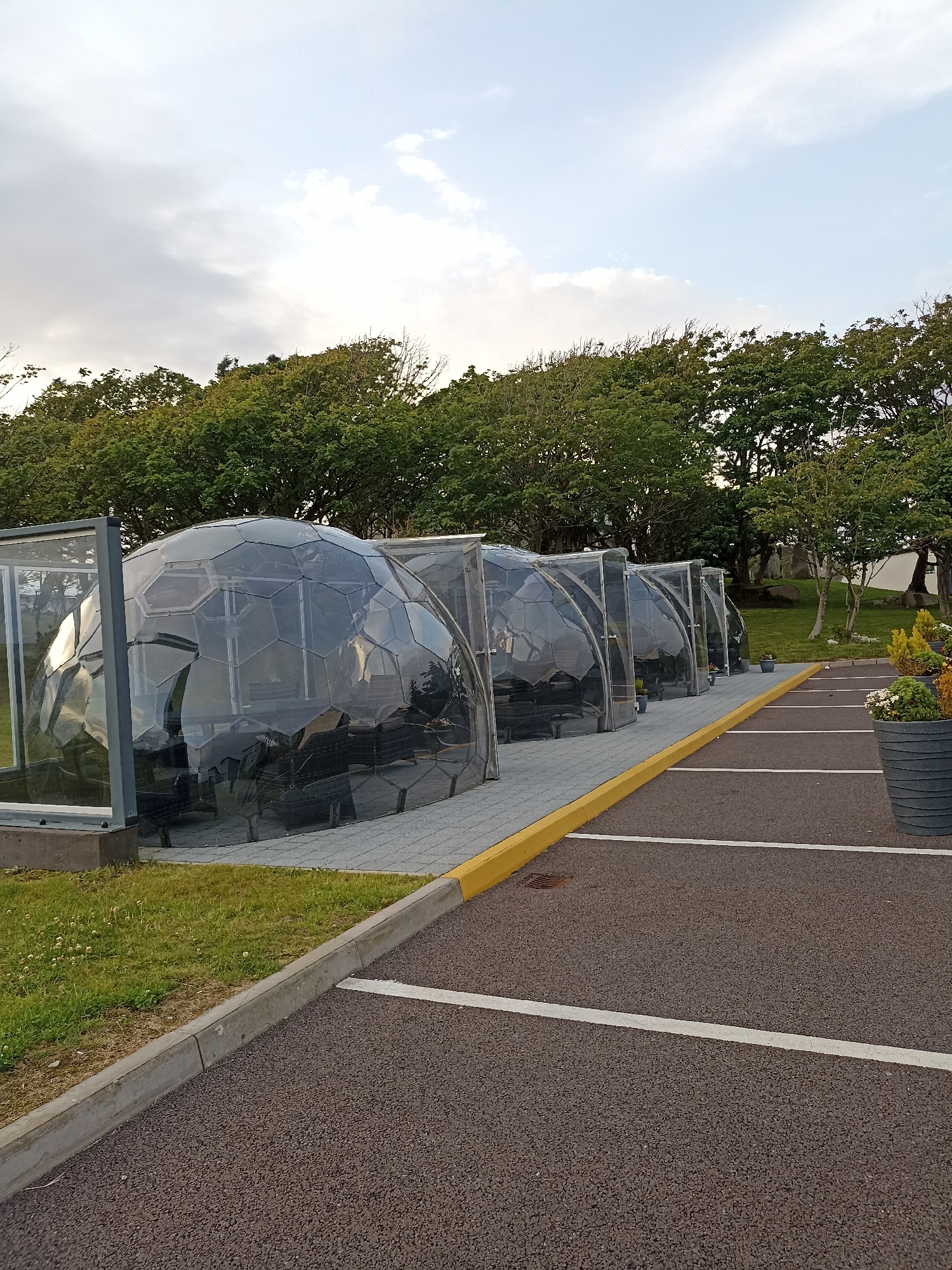 The image depicts a row of transparent geodesic domes set up in a parking lot. These domes appear to be individual dining or seating areas, each furnished with dark-colored seating. They are situated on a paved walkway bordered by a yellow curb and a grassy area. Beyond the domes, a line of trees forms the background, with a cloudy sky overhead. The parking lot itself is paved with dark asphalt and marked with white lines. In the foreground, to the right, a large dark gray planter contains various plants, including flowers and foliage. The scene is outdoors, under natural lighting conditions. The location is identified as Doirí Beaga, Ireland, suggesting this is an outdoor dining or recreational setup in that region. There are no people visible in the image.