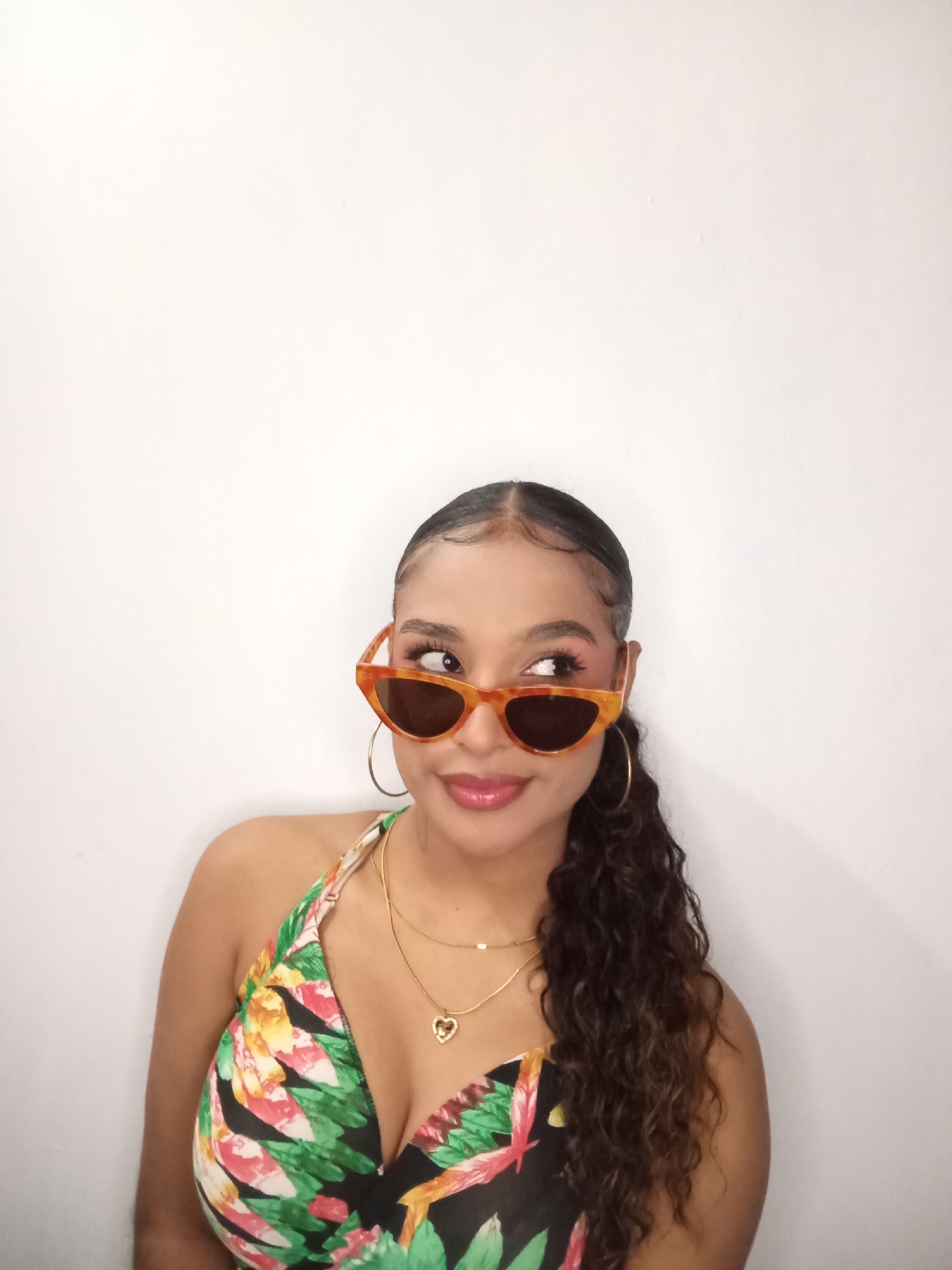 A young woman stands against a plain white wall. She wears sunglasses with a brown and orange frame and dark lenses, a halter top floral print swimsuit, gold hoop earrings, and two gold necklaces with a heart-shaped pendant. Her dark curly hair is pulled back. Her eyes are looking to the side. The location is not specified or implied in the image.