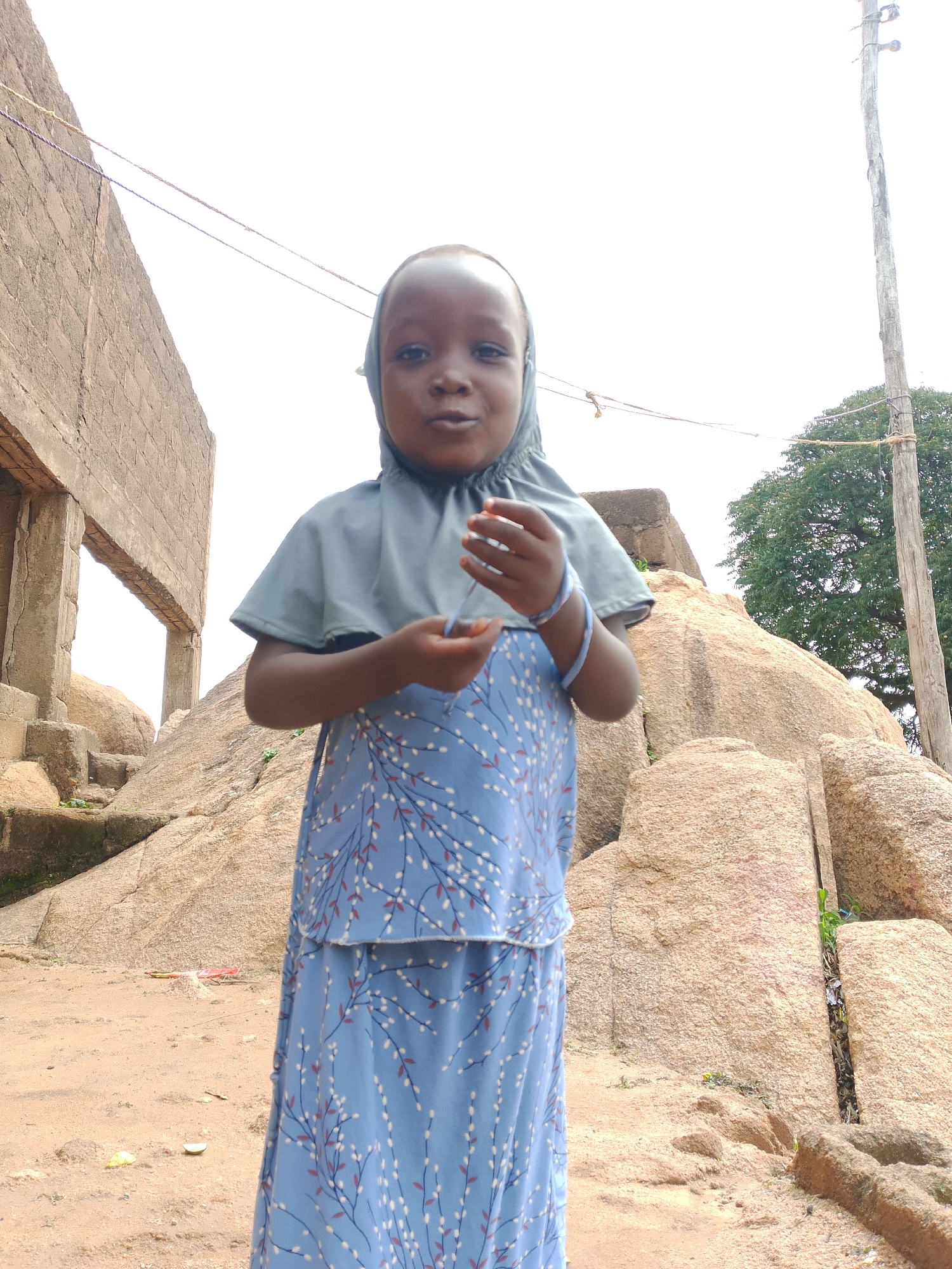 A young Black girl wearing a light blue hijab and a blue dress with a white and red floral pattern stands outdoors. She is looking at the camera and holding a strand of blue thread in her hands. Behind her is a large rock formation, a weathered concrete building, and a wooden utility pole with wires. The ground is sandy dirt.