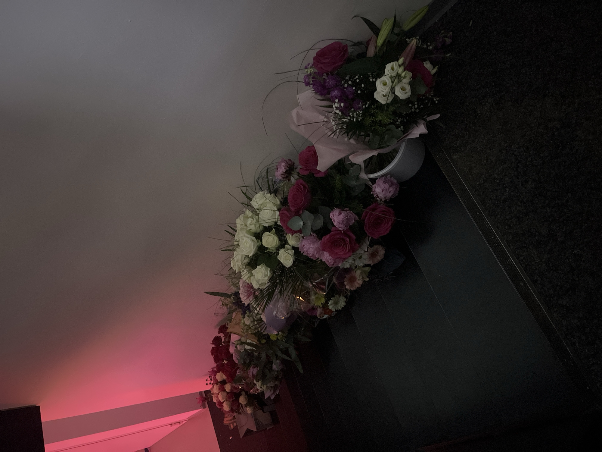 The image shows a floral arrangement displayed on a dark, possibly granite, surface. The arrangement is composed of various flowers, including roses, lilies, and greenery, in shades of white, pink, red, and purple. The flowers appear to be arranged in several containers, possibly vases or buckets, placed in a row. The background consists of a light gray wall and a section illuminated by a pink or red light. The scene appears to be indoors, possibly in a room or hallway. There is no indication of the image’s location in Saarbrücken, Germany. No people are visible.