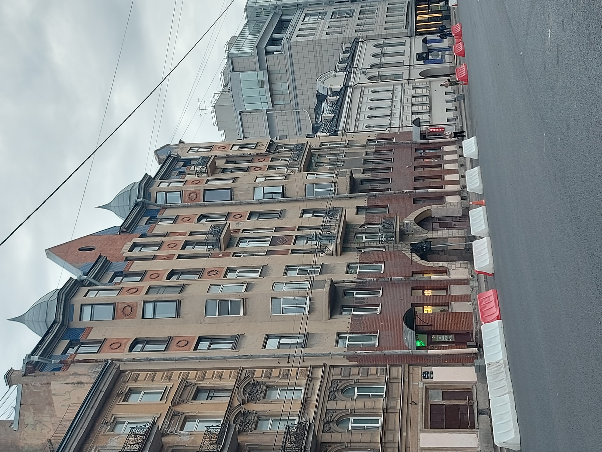 The image captures a street view in Saint Petersburg, Russia, showcasing a multi-story building with distinctive architectural features, including decorative patterns, rounded windows, and pointed roof elements. Adjacent to the building is a paved road with white and red barriers along the edge. Further down the street, modern glass and concrete buildings are visible. Several utility lines are strung across the sky. People walk near the barriers.