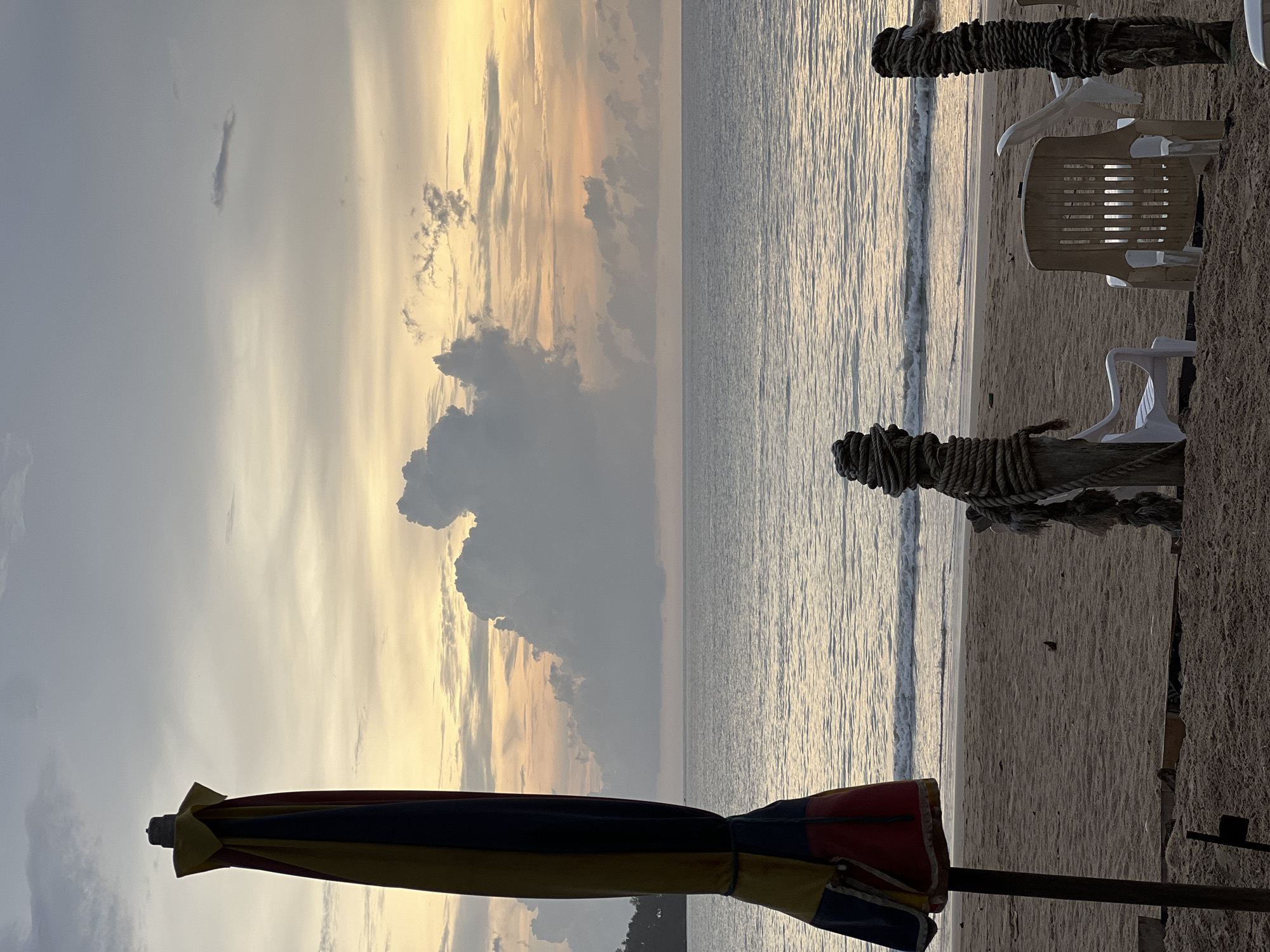 The image depicts a beach scene, likely at sunset, in an unknown location. The sky is filled with dramatic clouds, illuminated by a golden light. The sea is relatively calm with gentle waves. On the beach, there are beach chairs and a structure made of wood, possibly a support for a beach umbrella or shade. The edge of a partially folded umbrella is visible in the foreground, with a color scheme of yellow, blue, and red. The sand appears to be wet, suggesting recent high tide or wave activity. No people are immediately visible in the image.