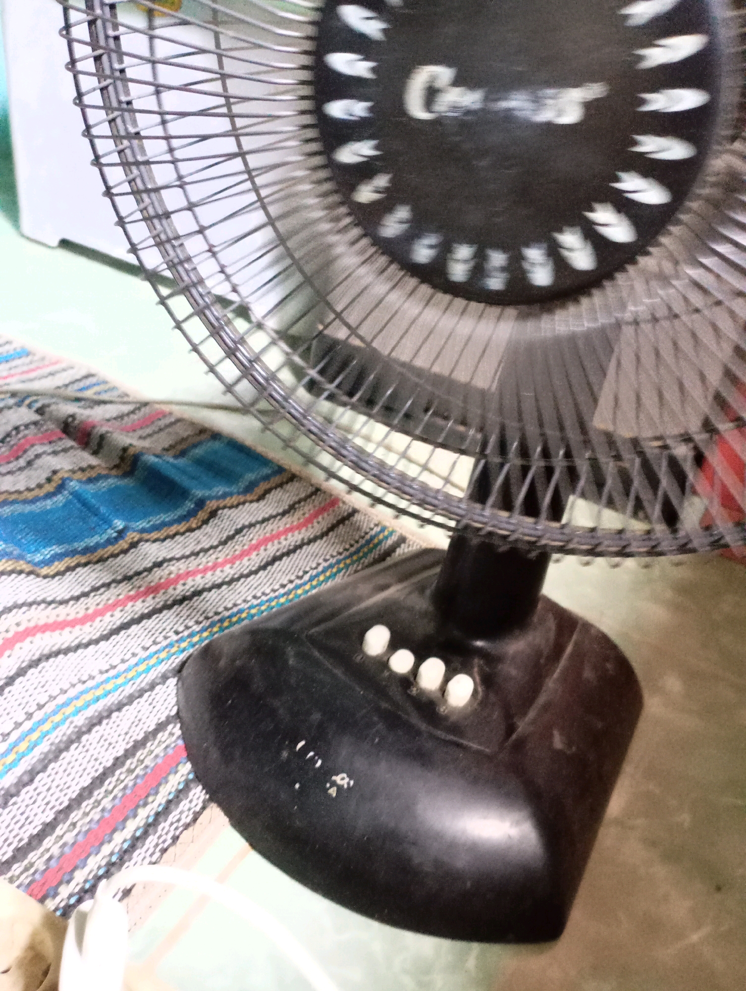 The image shows a close-up of a black electric fan, with its metal grill and part of its base visible. The fan is positioned on top of a colorful, woven mat. The mat has stripes of blue, white, red, and yellow. The fan's base has several white buttons. The background is blurred, but appears to be an indoor setting with a light green surface. The scene is static and does not depict any people or actions. No specific location in Kecamatan Haurgeulis, Indonesia can be determined from the visual information provided.