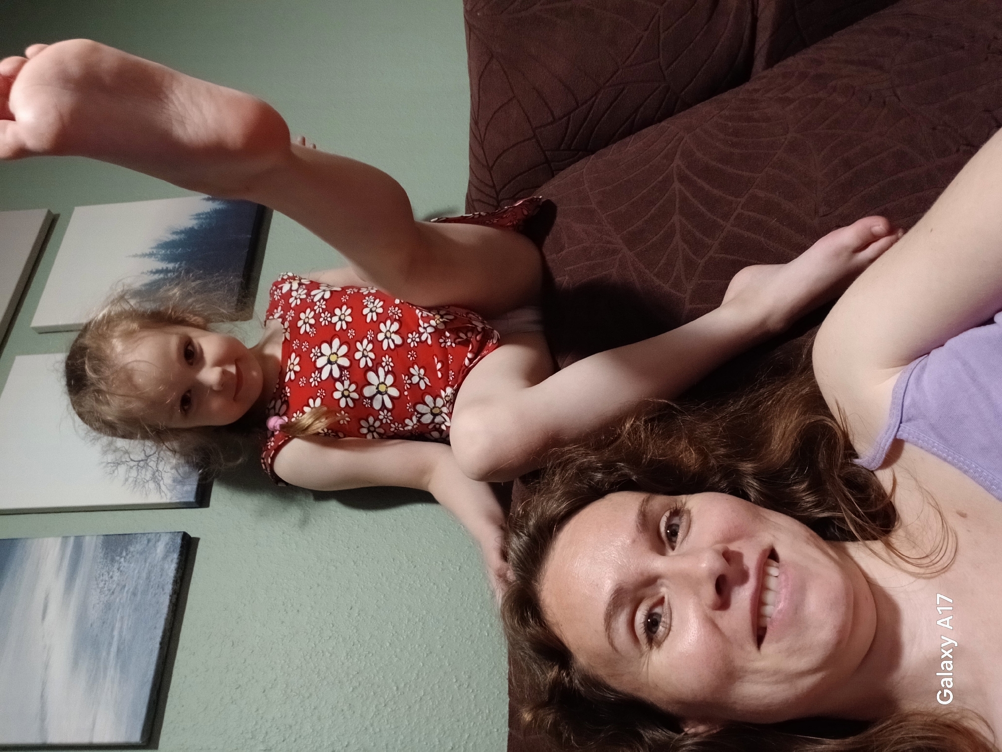 An adult female is visible in the foreground, smiling directly at the camera. She has brown, wavy hair and is wearing a light purple tank top. Behind her, a young female child with blonde, curly hair is seated on the backrest of a brown, textured sofa, also looking towards the camera with a slight smile. The child wears a red dress with white floral patterns. Her left leg is raised, exposing the sole of her foot and revealing white underwear beneath her dress. The background features a textured, light green wall adorned with two framed landscape artworks, one depicting a cloudy sky and the other bare trees. The image bears a "Galaxy A17" watermark in the lower-left corner. The scene is located in Yekaterinburg, Russia.