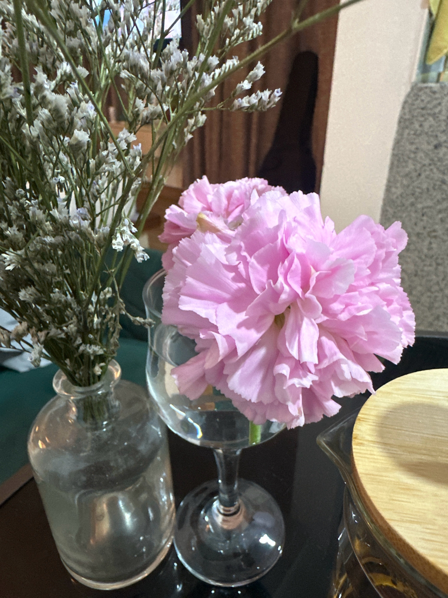 A close-up shot focuses on a pink carnation in a clear glass goblet, positioned to the right of a clear glass vase holding dried white flowers. The scene appears to be indoors, possibly a domestic setting, with a dark, reflective surface in the foreground. In the background, a blurred figure is visible, and part of a beige wall and a brown curtain can be discerned. There are no discernible Nepali cultural elements or geographical markers in the image that would definitively place it in Lalitpur, Nepal.