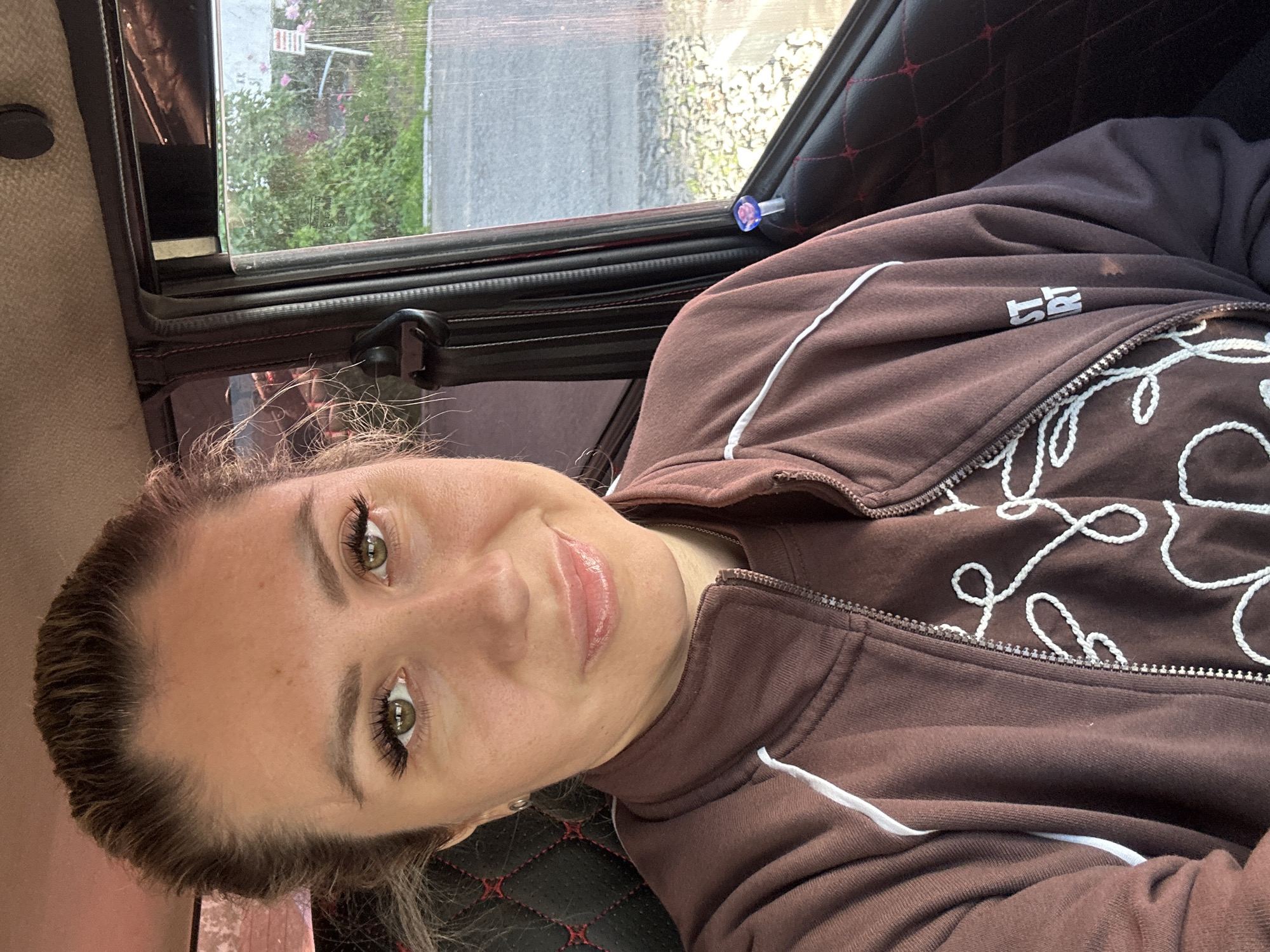 The image is a selfie taken by a woman inside a vehicle. She has medium-length brown hair pulled back in a ponytail, prominent eyelashes, and green eyes. She is wearing a brown zip-up jacket over a brown t-shirt with white lettering that reads "ST RT". The interior of the car features black and red quilted upholstery. Through the window, a glimpse of an outdoor environment is visible, including greenery, a paved area, and a sign in the distance. The weather appears to be overcast. The image was captured in Novoaltaysk, Russia.