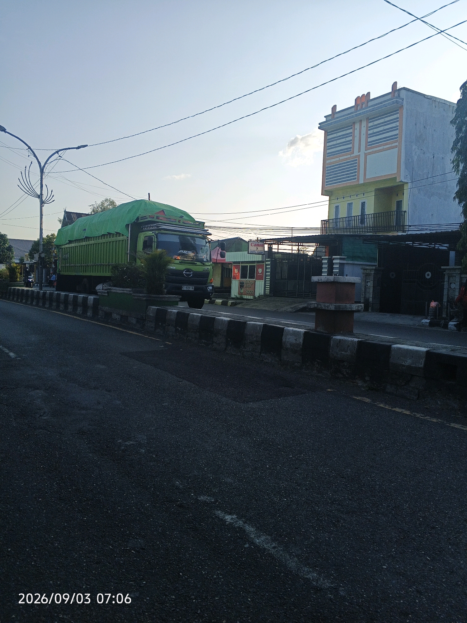 The image, timestamped 2026/09/03 07:06, captures a street scene in Pare-Pare, Indonesia. A green truck with a tarp-covered load is parked on the side of the road. The truck has a license plate reading "KT 8423 GL". Adjacent to the truck is a building with a yellow lower level and a gray upper level featuring a distinctive patterned facade with orange and blue accents. Further down the street, a lamppost stands prominently. The road has a black and white striped curb. In the background, there are various commercial establishments, including one with a "Canon" sign. The sky is clear with a hint of clouds.