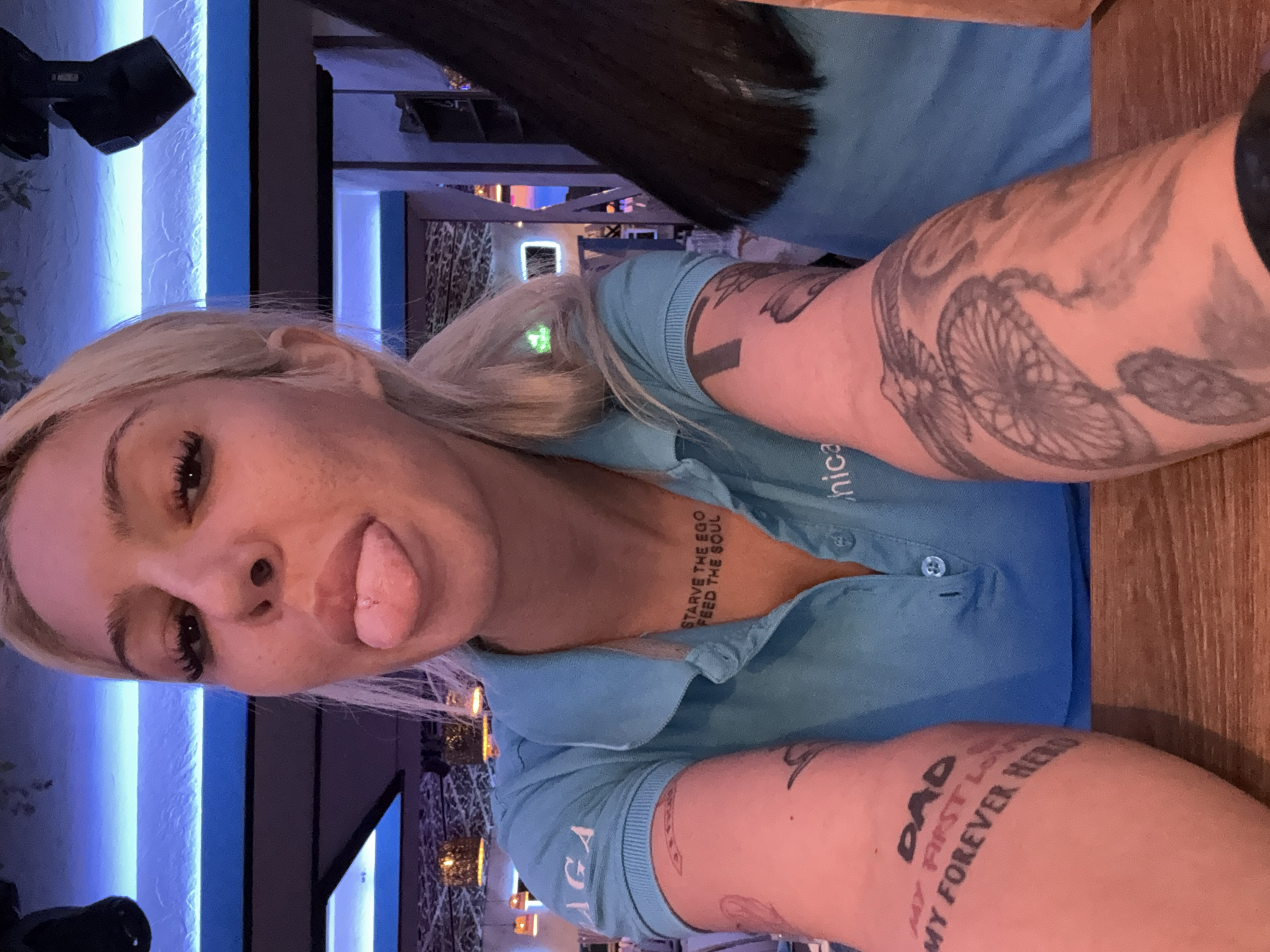 A close-up selfie captures a blonde woman with a playful expression, sticking her tongue out. She has dark eyes, prominent eyelashes, and a small nose piercing. Her long blonde hair is swept back, and a faint green light is visible near her ear, possibly a reflection. She is wearing a light blue collared polo shirt with ribbed cuffs; partial text such as "hnic" and "LAGA" is visible on the shirt, suggesting branded attire or a uniform.
The woman is heavily tattooed. On her chest, visible below her chin, is the text "STARVE THE EGO FEED THE SOUL". On her right forearm, a large, intricate black-and-grey tattoo features what appears to be a spoked wheel or gear, integrated into a larger design. Her left forearm displays the text "DAD MY FIRST LOVE MY FOREVER HERO".
The setting is an indoor establishment, likely a restaurant or bar in Plovdiv, Bulgaria, as indicated by the artificial lighting, table, and background elements. The dim ambiance suggests an evening or night-time scene. In the blurred background, vertical blue LED strips illuminate a textured wall, and warm-toned decorative hanging lights are visible. A black speaker or lighting fixture is mounted on a wall. A wooden table surface is prominent in the foreground, and a glimpse of another person's dark hair is visible in the upper background. The overall mood is informal and casual.