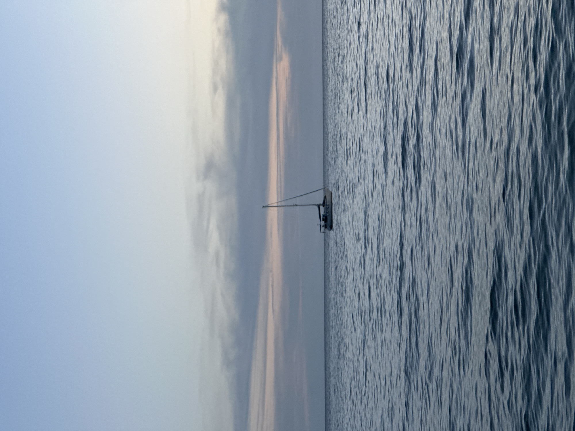 The image depicts a sailboat on the Adriatic Sea off the coast of Šušanj, Montenegro. The sky is a pale blue with streaks of gray and faint pink clouds near the horizon. The sea is slightly choppy with small waves and dark blue-gray in color. The sailboat is small and white, with its mast visible against the sky.