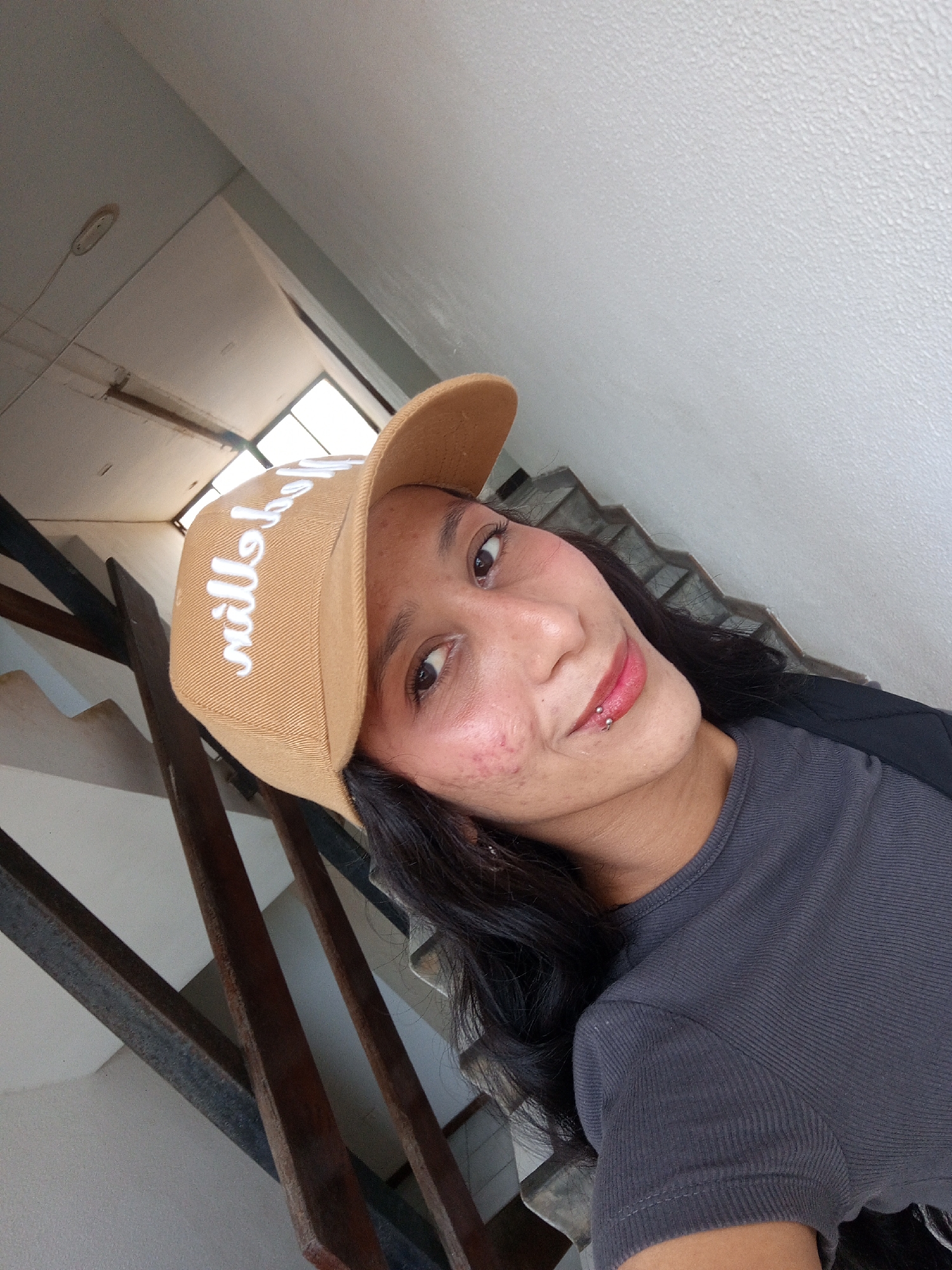 A selfie shows a woman with dark hair wearing a beige baseball cap, a gray t-shirt, and a lip piercing. She is smiling slightly. The background consists of a staircase with metal railings and a textured white wall. A window is visible at the top of the stairs, suggesting an interior location. The image appears to be taken inside a building in Porlamar, Venezuela.