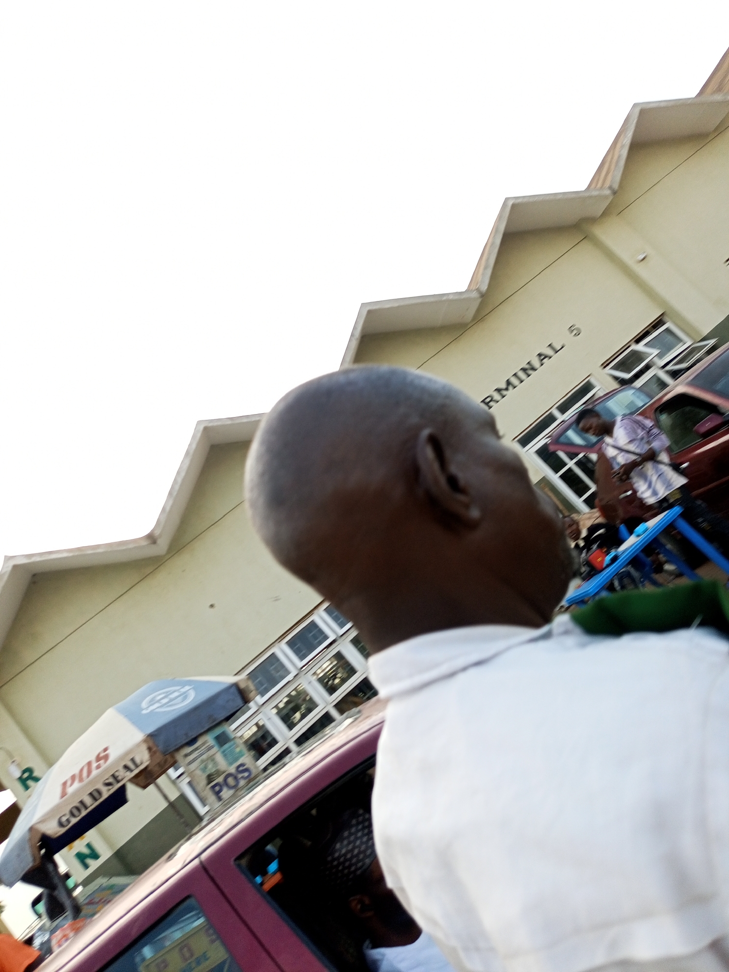The image depicts an outdoor scene, likely a busy area with a building in the background. The building appears to be a terminal, as suggested by the partially visible text "TERMINAL 5" on its facade. The architecture of the building is characterized by a stepped, angular design along the roofline. In the foreground, the back of a person's head and shoulder, wearing a white shirt, are visible, indicating the photographer is looking over their shoulder. Further into the scene, other individuals are present, including one person wearing a purple and white striped shirt, standing near a maroon car. A pinkish-purple car is also prominent, with an umbrella attached to its roof displaying "POS" and "GOLD SEAL." The overall environment suggests a public transportation hub or market area, possibly located in Gombe, Nigeria, given the context. The lighting indicates it is daytime, with a bright, overcast sky.