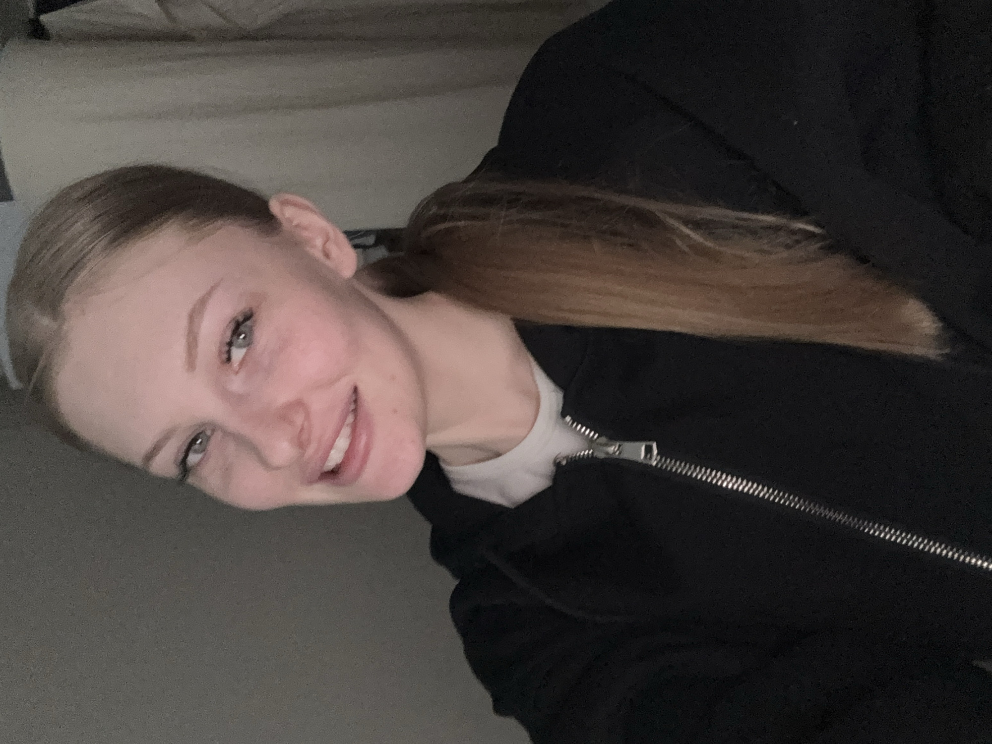 A young woman with fair skin, light brown hair pulled back in a ponytail, and wearing a black zip-up jacket over a white shirt is smiling at the camera. She is indoors, possibly at home, with a light-colored surface visible in the upper left corner, which could be a bed or couch. The image's orientation suggests it was taken from a slightly angled perspective, likely as a selfie. There is no information available to determine if the location is in the city of Perm, Russia.