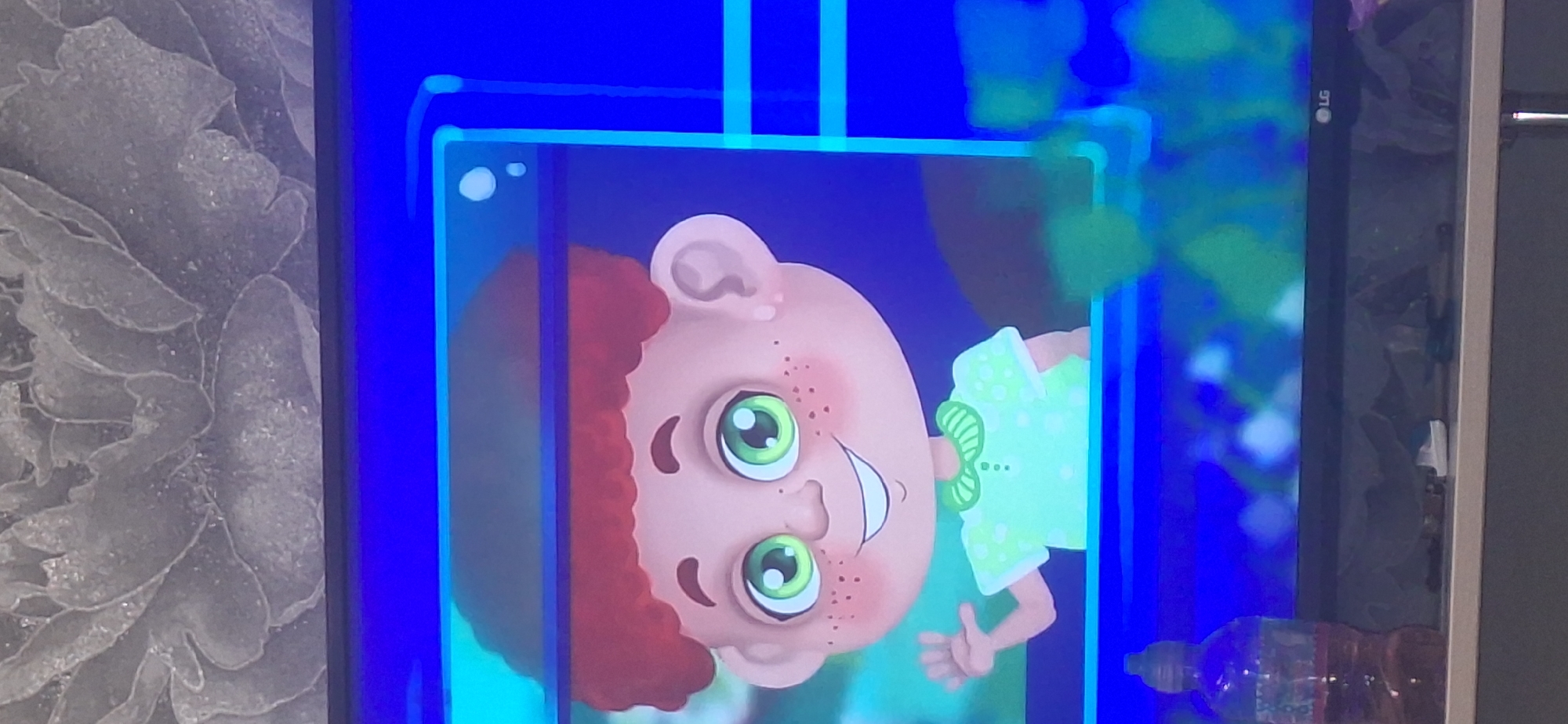 A television screen displays a cartoon of a child with red hair and green eyes, wearing a light green shirt with white polka dots. The child has freckles and is smiling, with one hand raised as if waving. The background is a vibrant blue with some abstract shapes and what appear to be bubbles. The television is placed on a shelf or surface, and in the foreground, a plastic bottle with a reddish liquid is visible. The overall scene is indoors, with the television being the primary focus. There is no information to determine the location in Bila Tserkva, Ukraine.