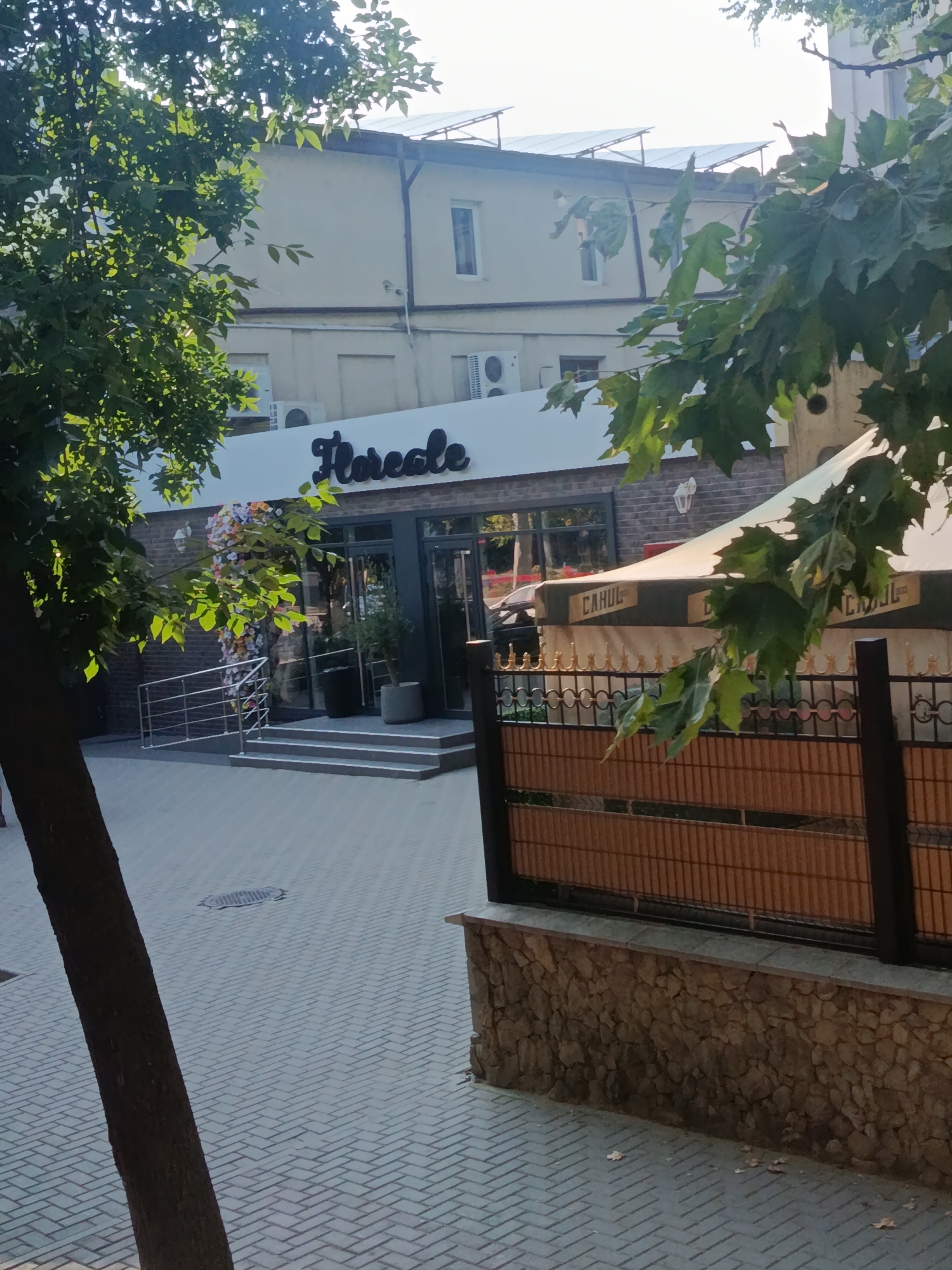 The image depicts the facade of a building with a prominent sign reading "Florcale" in a stylized font. The building appears to be a commercial establishment, possibly a cafe or restaurant, as suggested by the presence of outdoor seating under a large awning that reads "CAHUL". The awning itself is supported by a decorative metal fence. The building's exterior is made of brick and features large glass windows. In front of the building, there is a paved walkway with a tree in the foreground obscuring part of the view. Several steps lead up to the entrance of the building. The setting appears to be a city street in Cahul, Moldova, on a sunny day.