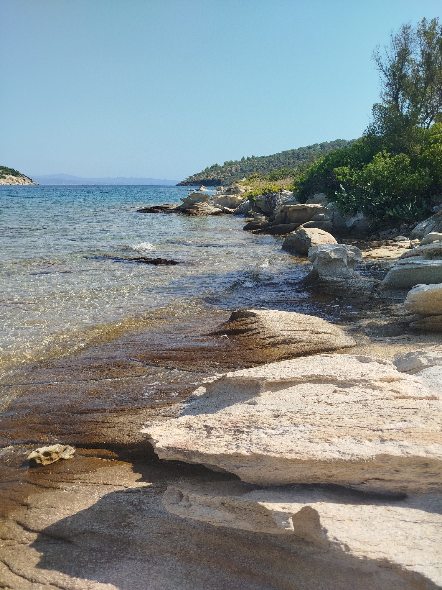 A bright, sunny day scene at a rocky coastline. The foreground is dominated by light-colored, textured rock formations, some layered and weathered, with shadows cast upon them. To the left, clear, shallow water gently laps over submerged rocks and sandy patches, showing ripples and sunlight reflecting on the surface. Further out, the water is a deeper blue, meeting a hazy, distant shoreline with green hills. The middle ground features more rocks and vegetation, including trees and shrubs, lining the coast. Two small boats are visible anchored offshore. The sky is a clear, pale blue. The location is a beach in Sithonia Municipal Unit, Greece.