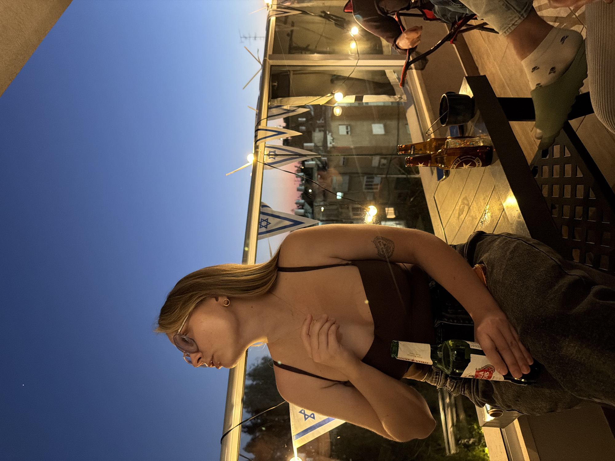 The image captures a relaxed evening gathering on what appears to be a balcony or outdoor patio, overlooking an urban landscape at twilight. The deep blue sky is visible on the left, contrasting with the warm, artificial lighting of the setting.
A woman with long, light brown hair and glasses is prominently featured in the foreground, seated and looking towards the left. She wears a dark brown spaghetti-strap top and has a tattoo on her upper left arm. She holds a green bottle of Stella Artois beer in her right hand, while her left hand rests near her chest. Nearby, parts of other individuals are visible, including a leg wearing a light green sandal and a white sock with small patterns, and an arm with a red strap.
The outdoor space is decorated with a string of small Israeli flags, running along what seems to be a glass barrier or window that separates the area from the city below. Bright string lights illuminate the immediate surroundings, casting a warm glow. Through the glass, the faint outlines of residential buildings and an antenna are discernible in the distance. On a table in the background, two amber-colored bottles and a black mug can be seen.
The visible text "STELLA ARTOIS" is clearly legible on the green bottle the woman is holding. The scene suggests a casual social event or celebration taking place in Ramat Gan, Israel, as indicated by the flags and the urban context.