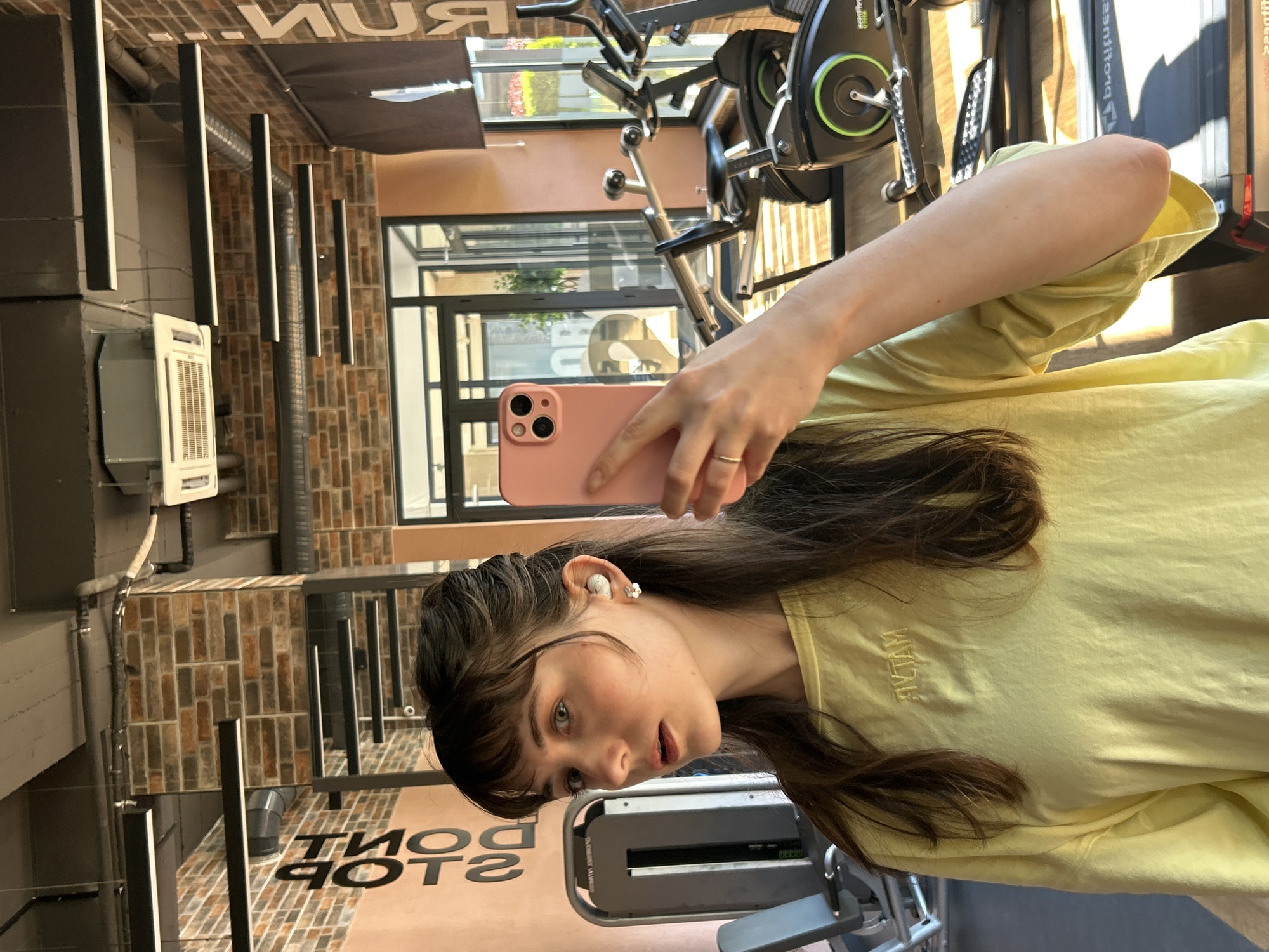 A person is taking a selfie in a gym, likely in Alanya, Turkey. The person is wearing a yellow t-shirt and has brown hair with bangs. They are holding a pink smartphone. The gym contains exercise equipment like treadmills and stationary bikes. The interior design includes brick walls, black metal fixtures, and a light-colored floor with a motivational message. Through the windows, one can see outdoor greenery and the city environment.