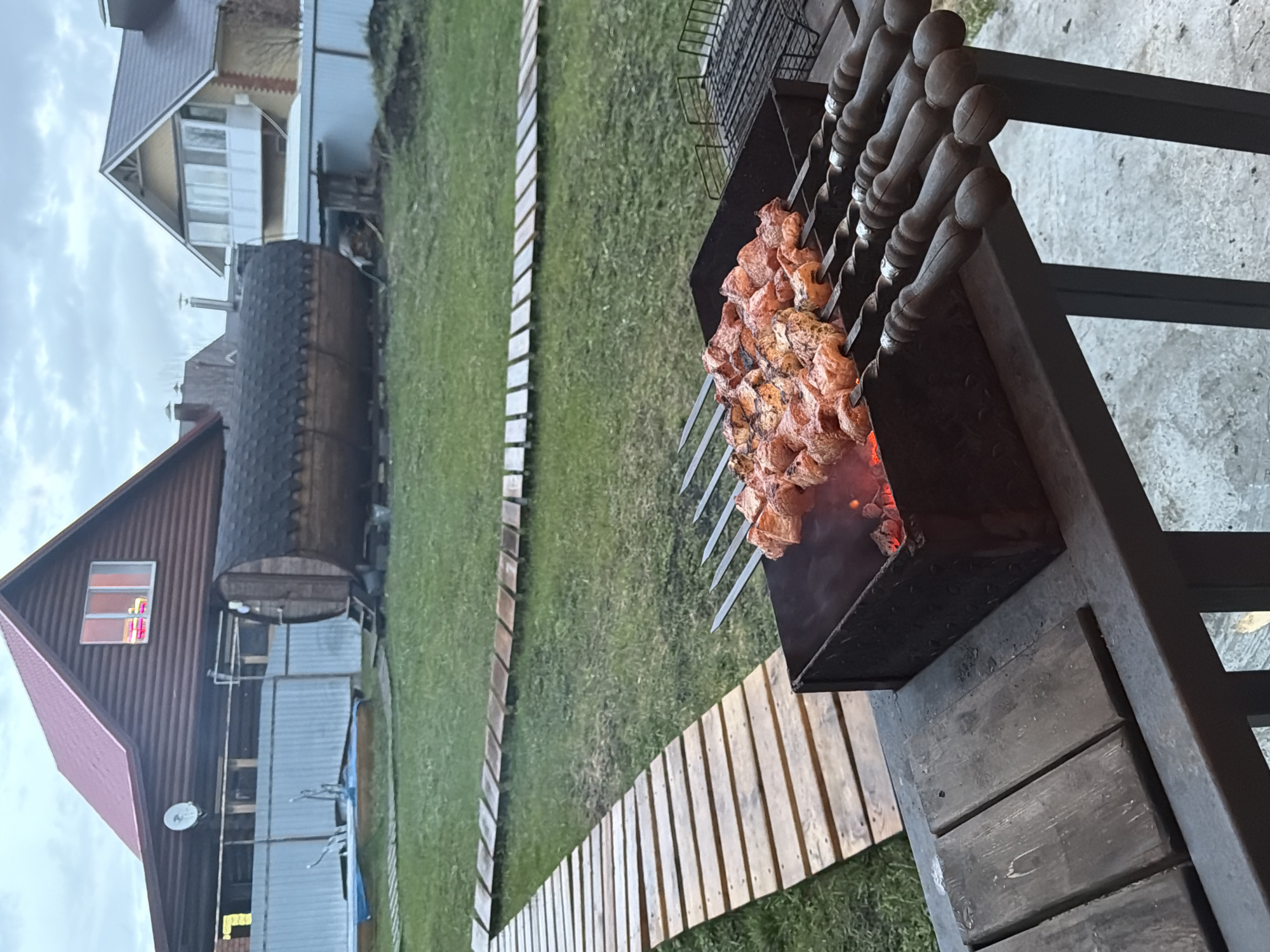 The image shows a barbecue taking place in Большая Шильна, Russia. Several skewers of meat are arranged over hot coals in a black metal grill. The skewers have wooden handles. The grill is placed on a wooden structure. To the left, there is a grassy lawn with a wooden pathway leading to a house with a red roof and a wooden building next to it. The sky is overcast.