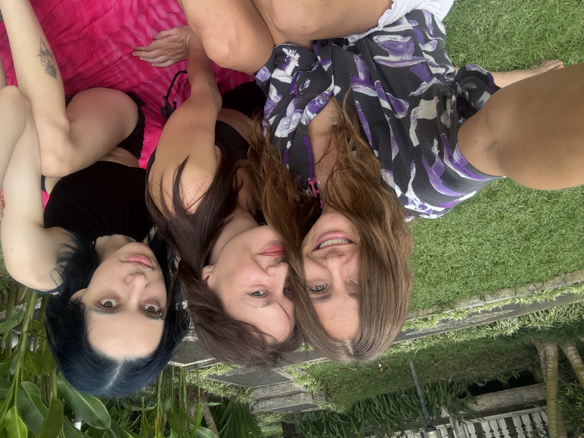 Three women are lying down in an outdoor setting. The woman on the left has dark blue hair and is wearing a black top. The woman in the middle has dark brown hair and is also wearing a black top. The woman on the right has light brown hair and is wearing a patterned black and purple shirt. The woman on the left is lying on a pink blanket, while the other two women are lying on grass. The background includes plants and a white railing. The scene appears to be located in Mengwi, Indonesia.