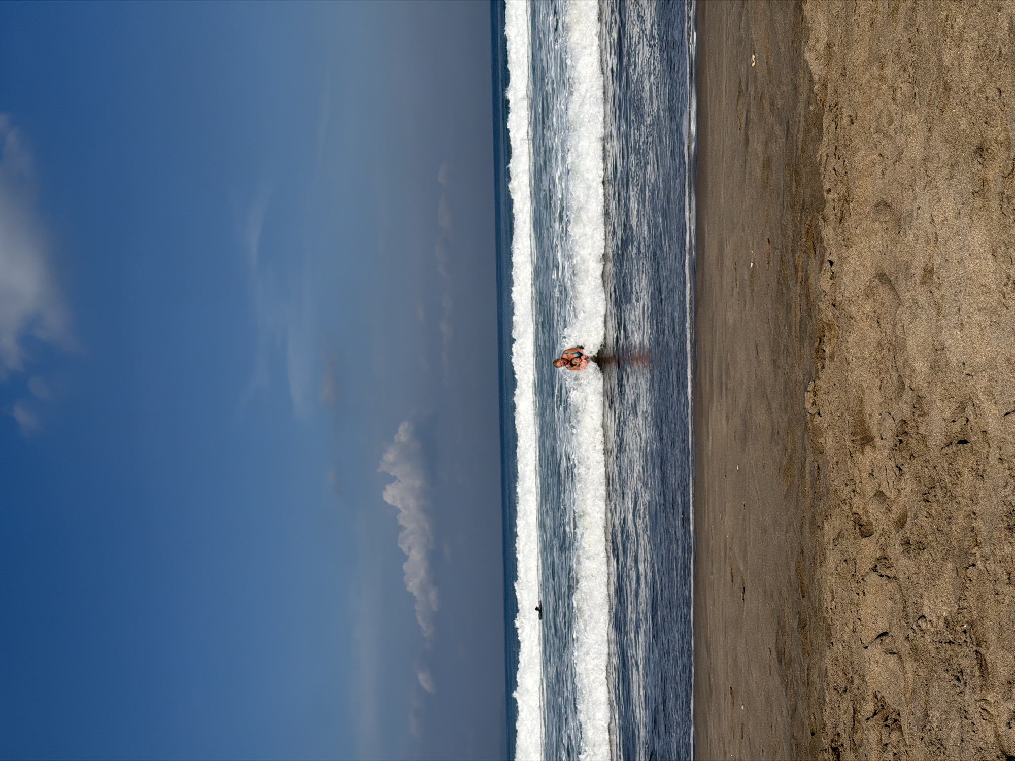 The image displays a serene beach scene, likely Kuta Beach in Indonesia, under a bright, clear blue sky. The composition is horizontally divided into three main sections: the vast sky at the top, the ocean with waves in the middle, and the sandy beach at the bottom.
The sky is a vibrant blue, featuring scattered white clouds that add depth and suggest good weather conditions. The ocean is characterized by multiple lines of white-crested waves actively breaking and rolling towards the shore. In the mid-ground of the ocean, a person wearing swimwear is visible, partially submerged in a breaking wave, suggesting they are swimming or bodyboarding. A smaller, darker figure can be faintly seen further out towards the horizon, possibly another person in the water.
The sandy beach, occupying the lower third of the frame, is light brown and appears somewhat coarse, with visible textures and minor imperfections. Footprints and small debris are discernible on the sand, indicating recent activity. The overall impression is that of a sunny, pleasant day, likely mid-day, at a popular beach, with individuals enjoying the ocean. No visible text is present in the image.
