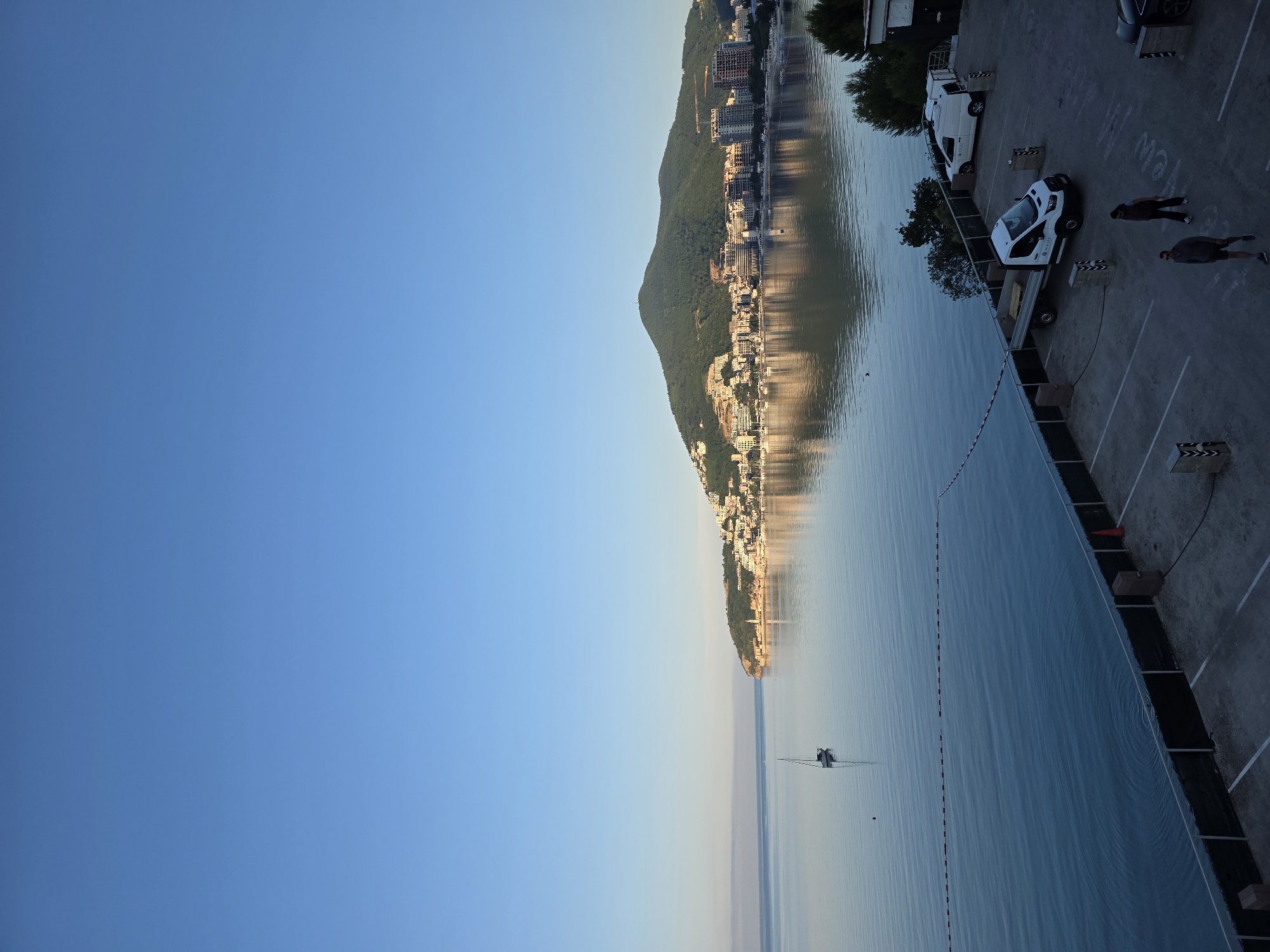 The image depicts a serene coastal scene in Budva, Montenegro, at the base of a lush, green mountain that slopes down to the sea. The mountain is dotted with white buildings, suggesting a densely populated area. The sea is calm and reflects the light from the sky, which is a clear, pale blue, indicative of early morning or late afternoon. A lone sailboat is visible on the water, adding a touch of tranquility. In the foreground, a parking lot is laid out on a paved surface, marked with white lines. Several vehicles are present, including a white van and a white flatbed truck with wooden pallets in its bed. Two individuals are walking through the parking lot, one wearing a grey hoodie and shorts, and the other in dark clothing. Another parked car is partially visible on the right side of the frame. A black barrier runs along the edge of the parking lot, separating it from the water, and a red and white striped rope is stretched across the water near the barrier, possibly marking a swimming area or a boundary. The overall impression is one of a peaceful morning or evening by the Adriatic Sea.