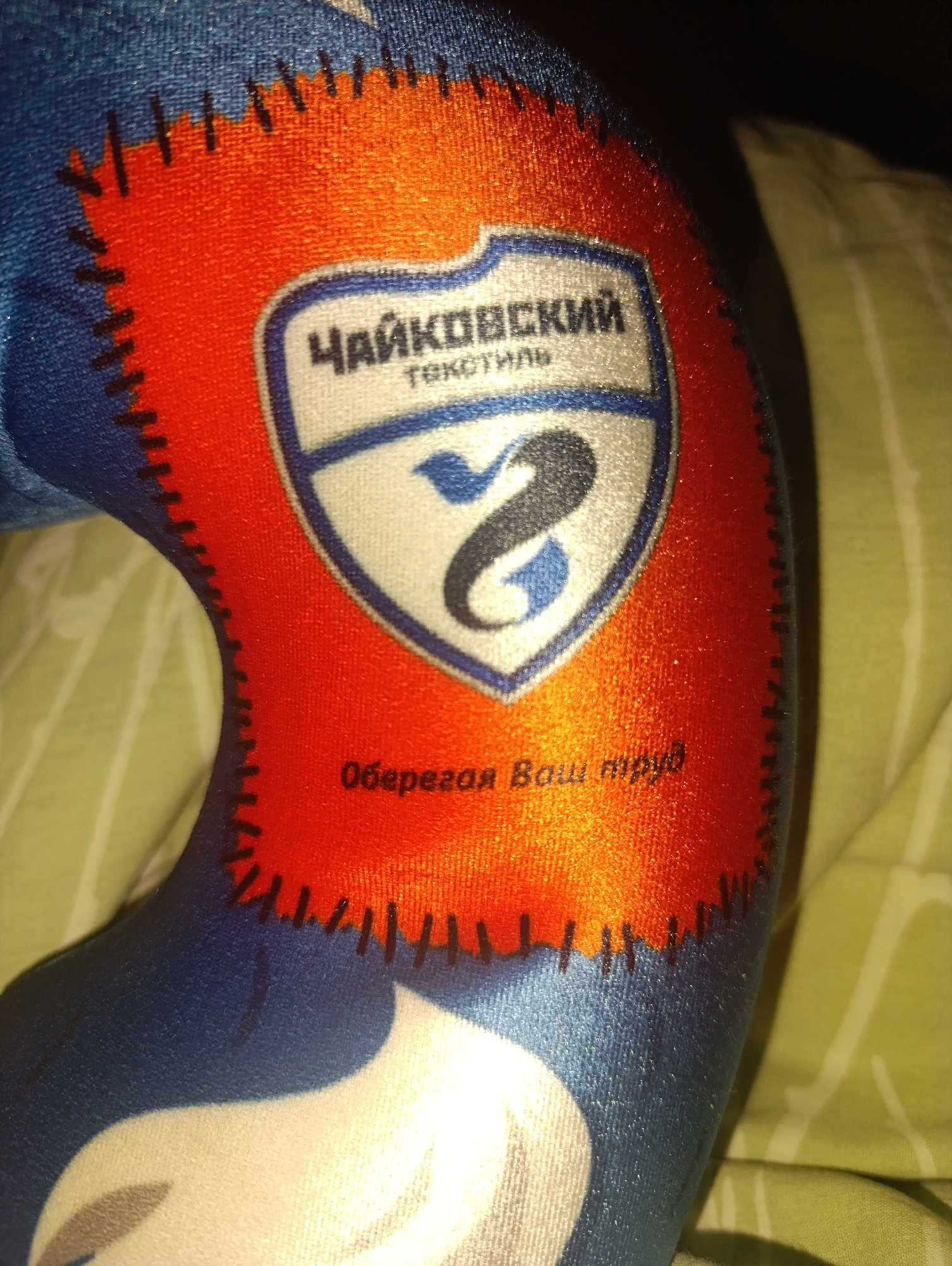 The image shows a neck pillow with an orange patch. The patch is bordered with black stitching and features a white crest with blue accents. The crest contains the text "Чайковский текстиль" and a stylized symbol. Below the crest is the text "Оберегая Ваш труд". The pillow is resting on a green and white striped surface. The image is brightly lit. There is no information about the location Зипуново in the image.