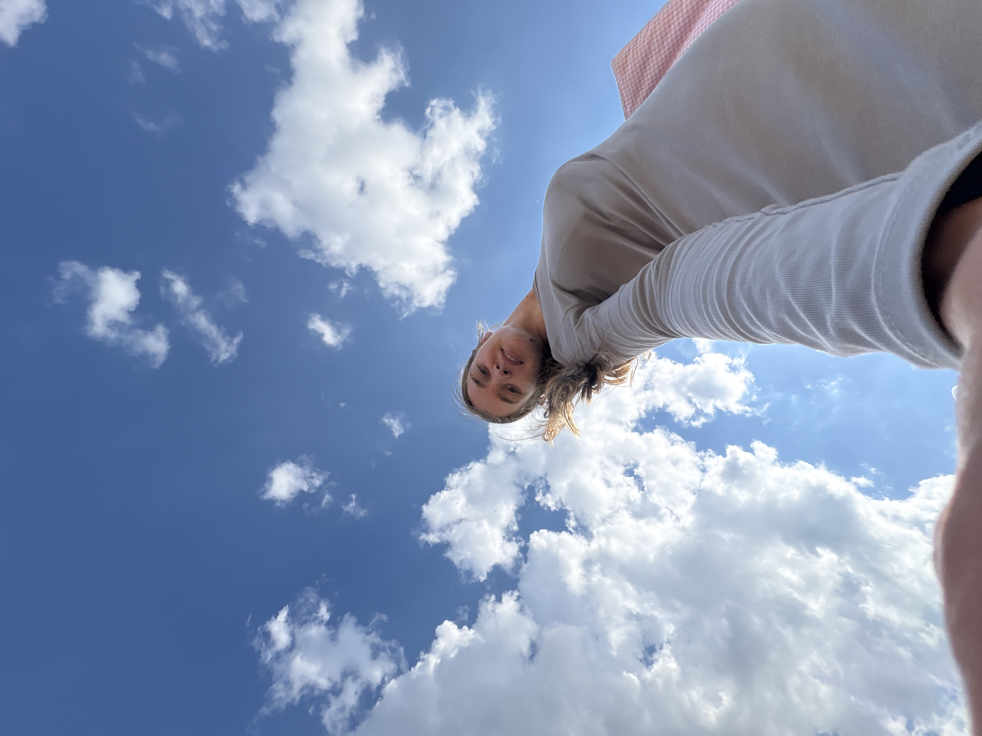 An upward-facing, low-angle, outdoor, self-portrait features a young Caucasian woman against a bright blue sky dotted with fluffy white clouds. She is wearing a long-sleeved, taupe-colored dress and her hair is a light brown color. Her head is tilted back, and she is looking towards the camera with a slight smile. Her left arm is extended towards the camera, suggesting she is holding it. To the right side of the frame, a portion of a pink and white checkered bag is visible. The overall impression is that of a person enjoying a sunny day outdoors. There are no discernable urban or natural landmarks that would allow for specific identification of the location within Prikamskii, Russia.
