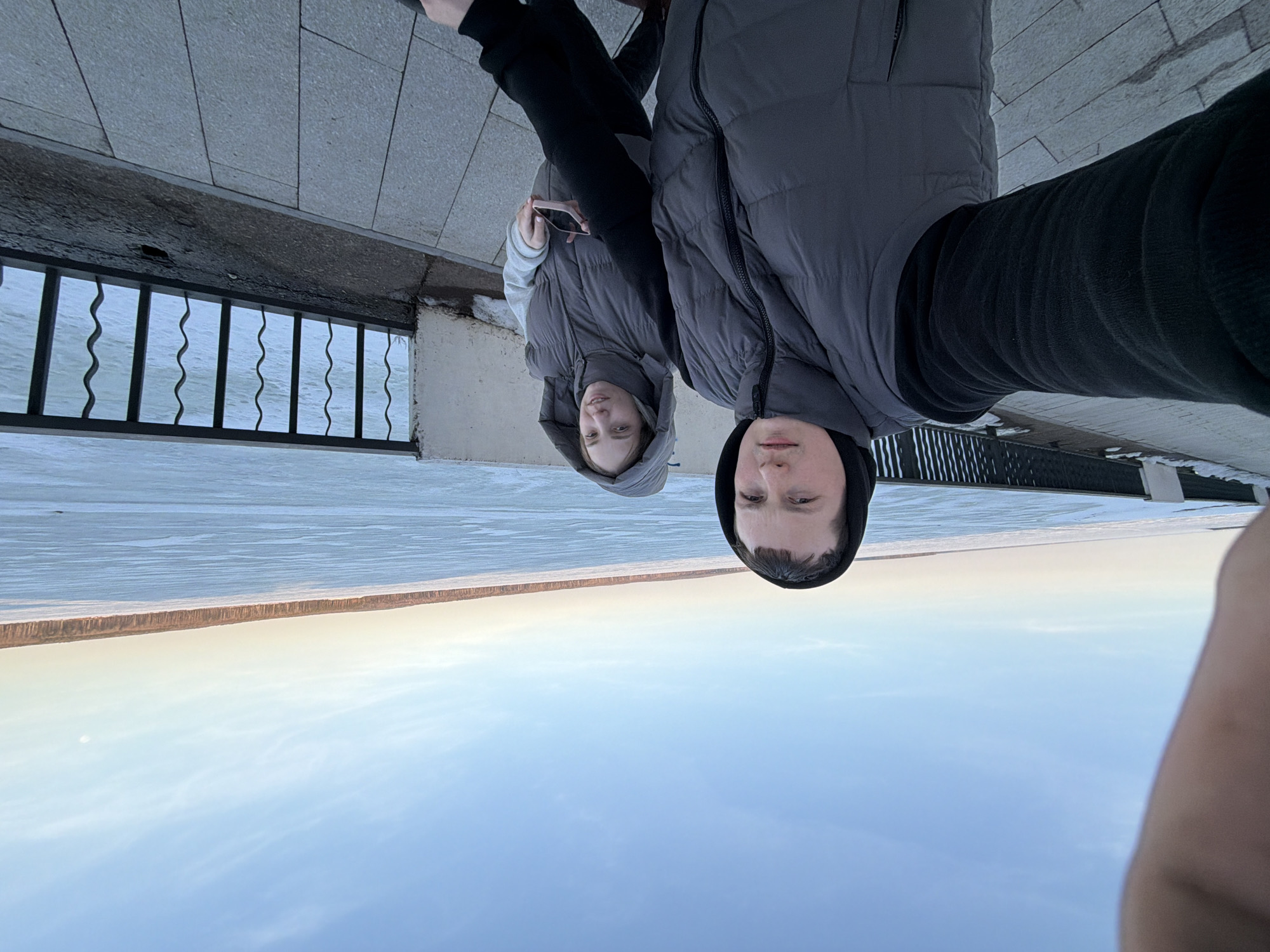 Two individuals are positioned in front of what appears to be a partially frozen body of water. The image is taken from an unusual, almost upside-down angle. One person is wearing a dark hoodie and a gray puffer vest, while the other is in a lighter gray hooded jacket and vest. A railing and a concrete edge are visible behind them. The backdrop features a horizon line with what may be trees or structures in the distance, under a sky with a slight blue tint. The setting is likely the city of Chaikovsky, Russia, based on the location referenced.
