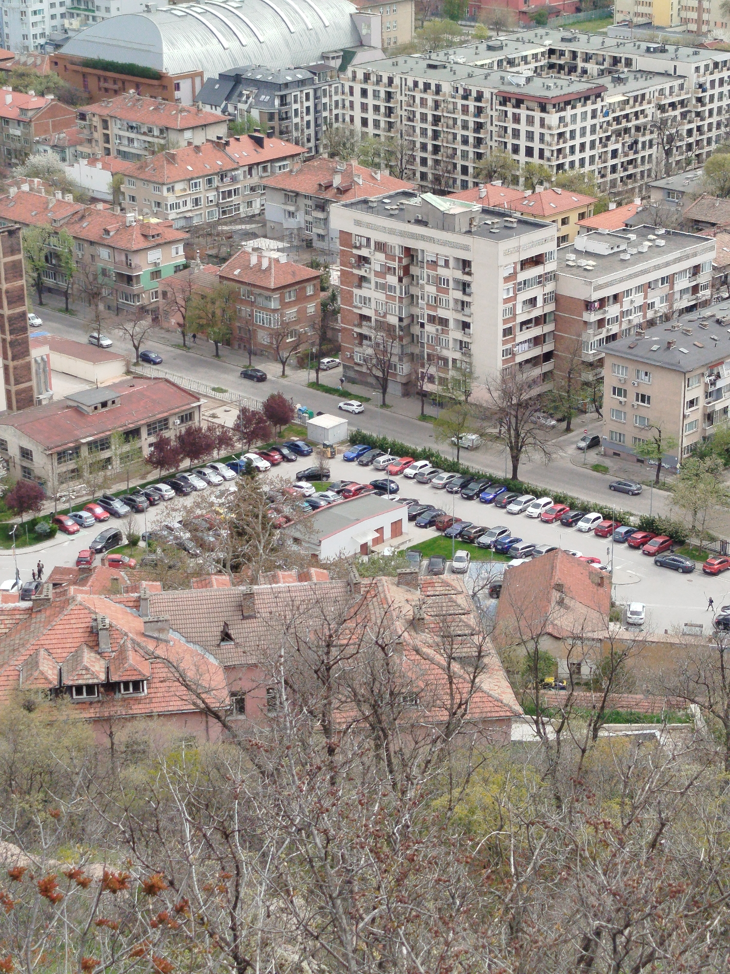 This is an elevated, aerial shot of a residential area in Plovdiv, Bulgaria. In the foreground, there are trees with bare branches, suggesting a late autumn or early spring season. Below the trees are buildings with red-tiled roofs. A parking lot is visible behind the buildings, filled with a variety of cars. Behind the parking lot, a few medium-rise buildings are arranged in rows. In the background, there is a large, silver-roofed building with curved top, surrounded by other apartment buildings. Cars are parked on streets and the weather looks overcast. No people are clearly discernible in the wide aerial view.