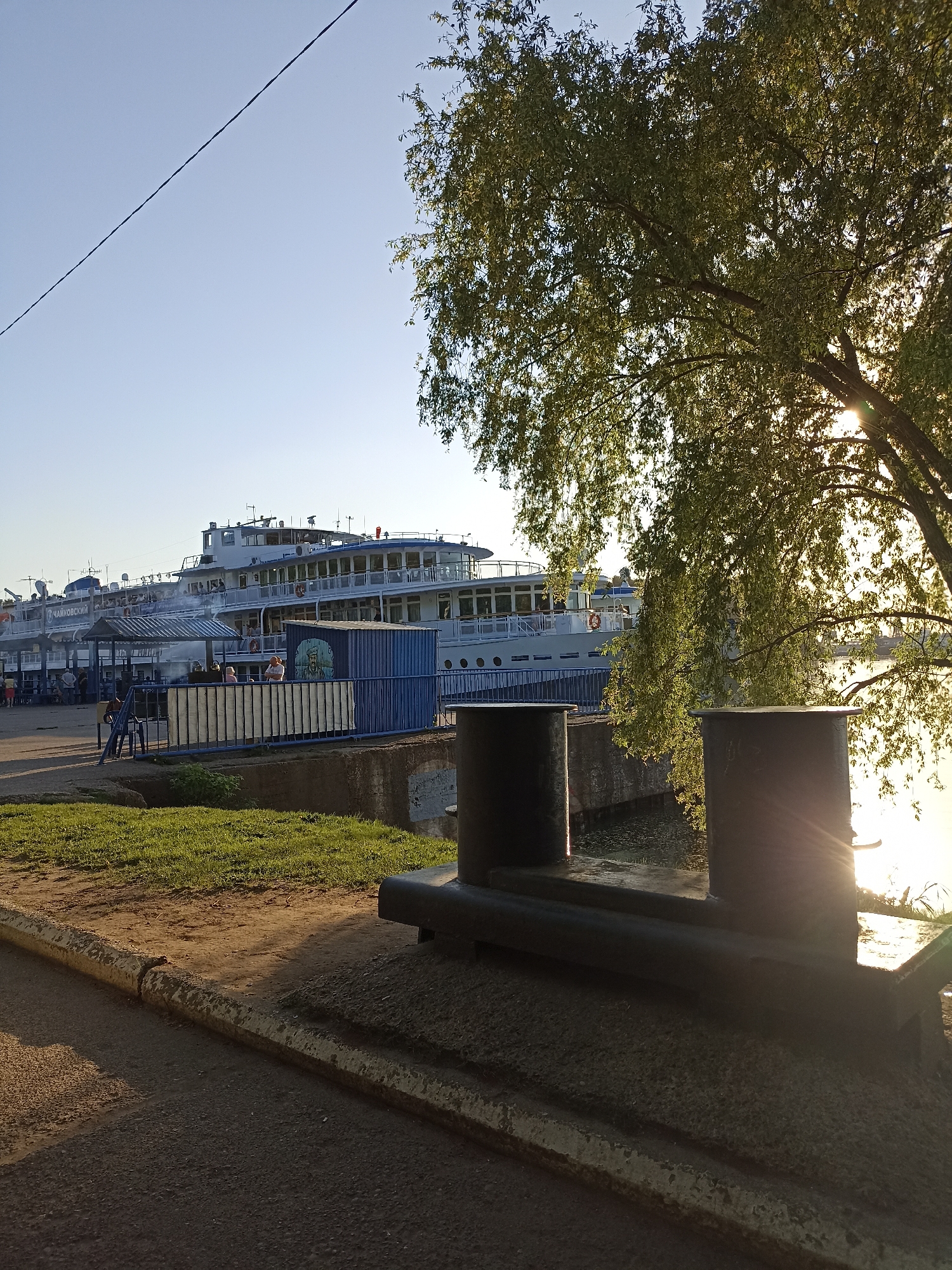 The image shows a three-tiered cruise ship docked at a pier. The ship has a white exterior with multiple windows and balconies. People are visible on the ship and on the pier. In the foreground, there are two black bollards connected by a horizontal bar, set on a concrete base. Grass and a paved walkway lead to the water's edge, where the bollards are located. A tree with green leaves partially obscures the view of the water and the sun. The sky is clear and blue. An powerline passes diagonally across the upper left of the frame.