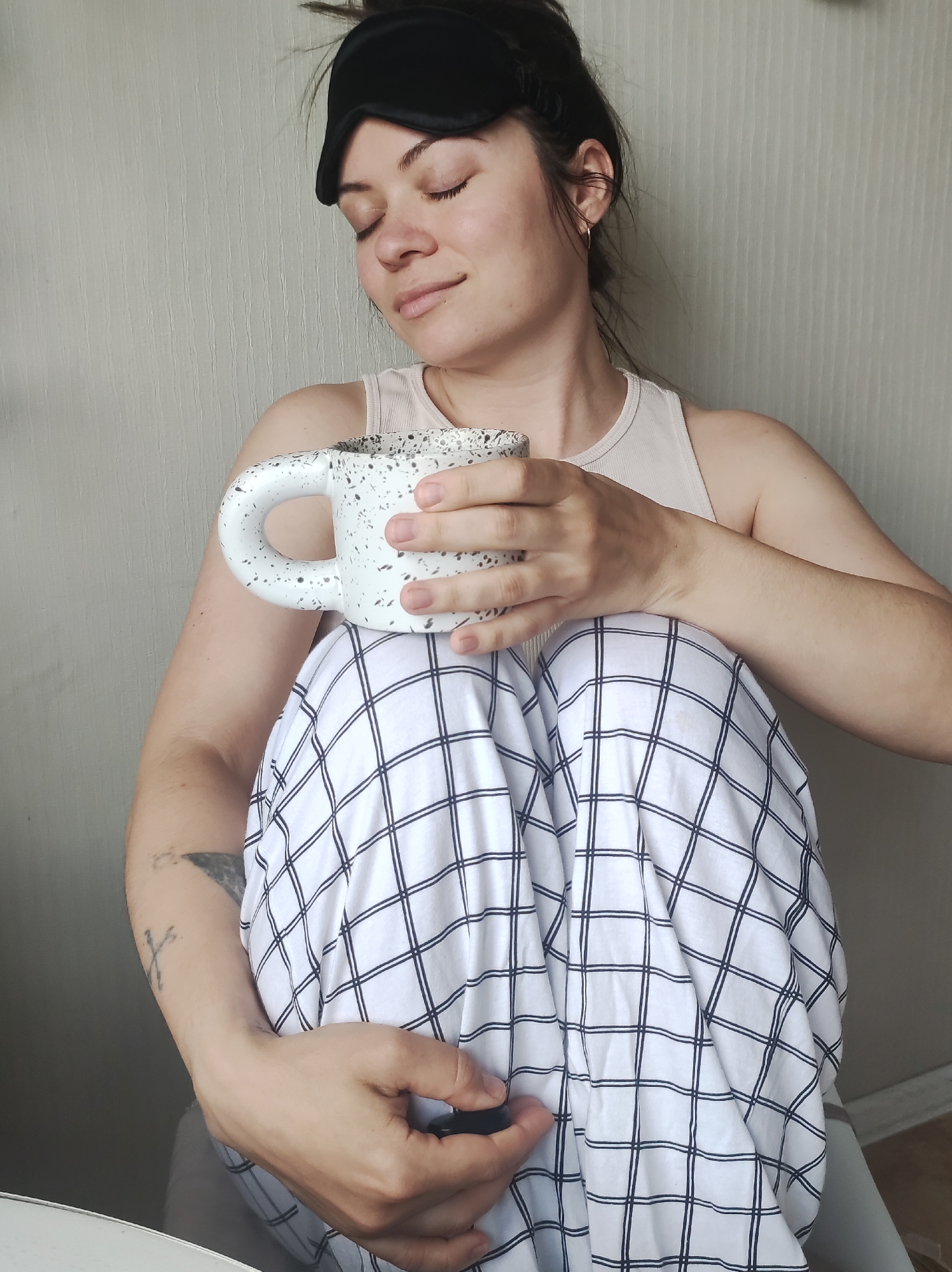A young woman is sitting indoors, holding a white mug with black speckles. She is wearing a black sleep mask on her forehead and has her eyes closed with a serene expression. She is wearing a light-colored tank top and white pants with a black grid pattern. Her arms are wrapped around her legs. The background is a plain beige wall.
There is no information about this photo being taken in Chaikovsky, Russia.