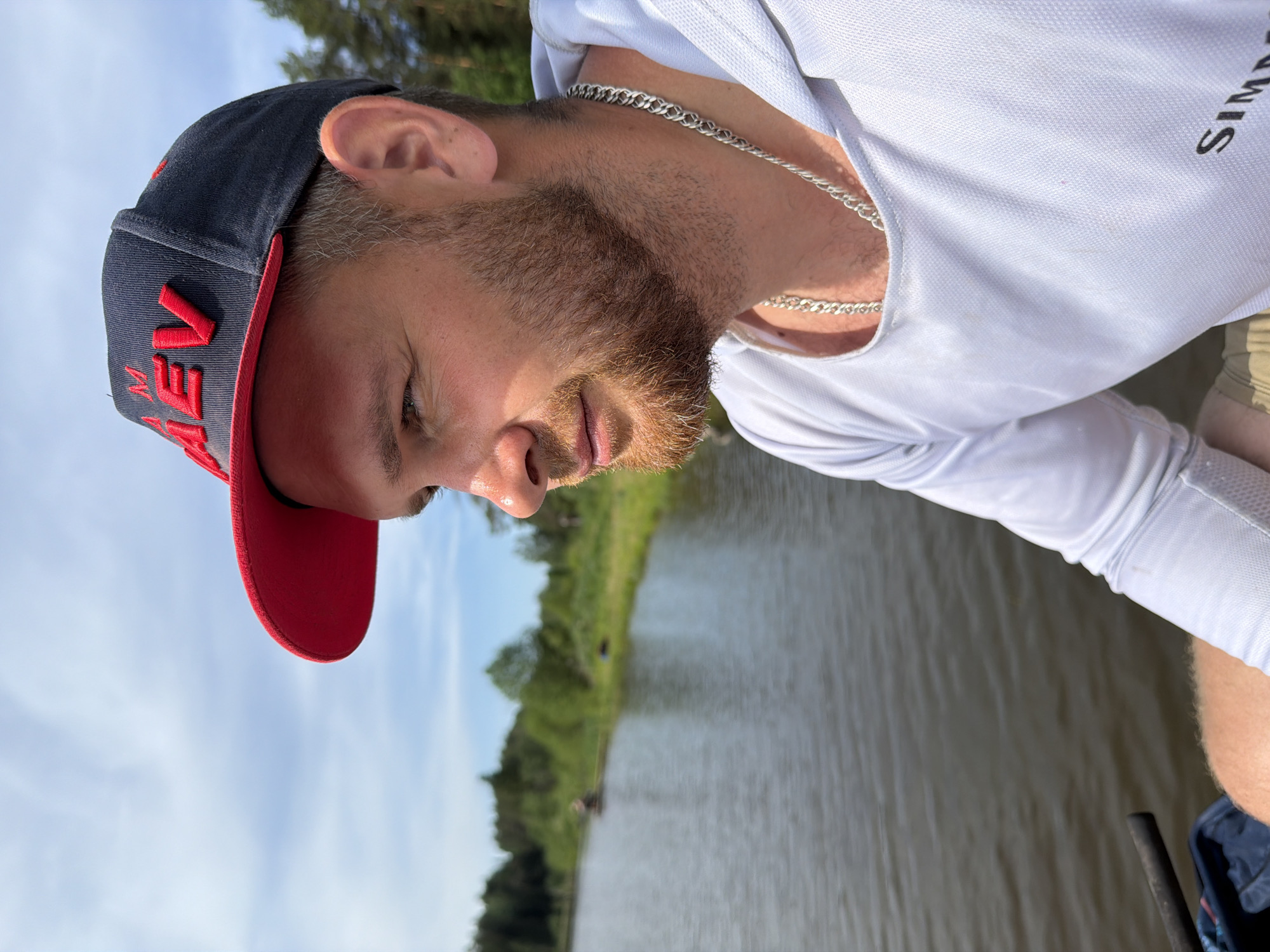 A man with a beard is shown in an outdoor setting. He is wearing a white long-sleeved shirt and a baseball cap with a red brim. A silver chain is visible around his neck. The background features a body of water, trees, and a sky with some clouds. The location is likely Prikamskii, Russia, based on the image prompt.