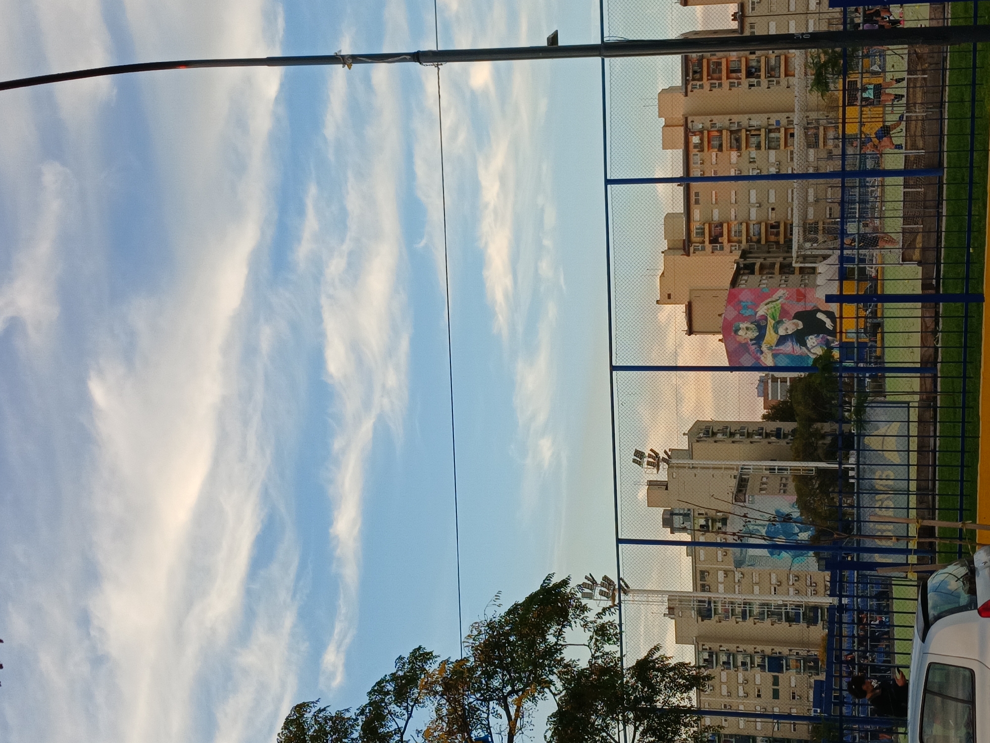 The image shows a sports field in Buenos Aires, Argentina, with apartment buildings in the background. A fence surrounds the field, and a mural of Lionel Messi is visible on one of the walls. There is a blue sky with streaks of clouds. Trees are visible in the foreground, and a utility pole is present. The image is taken from a low angle. A white car can be seen in the lower right corner.