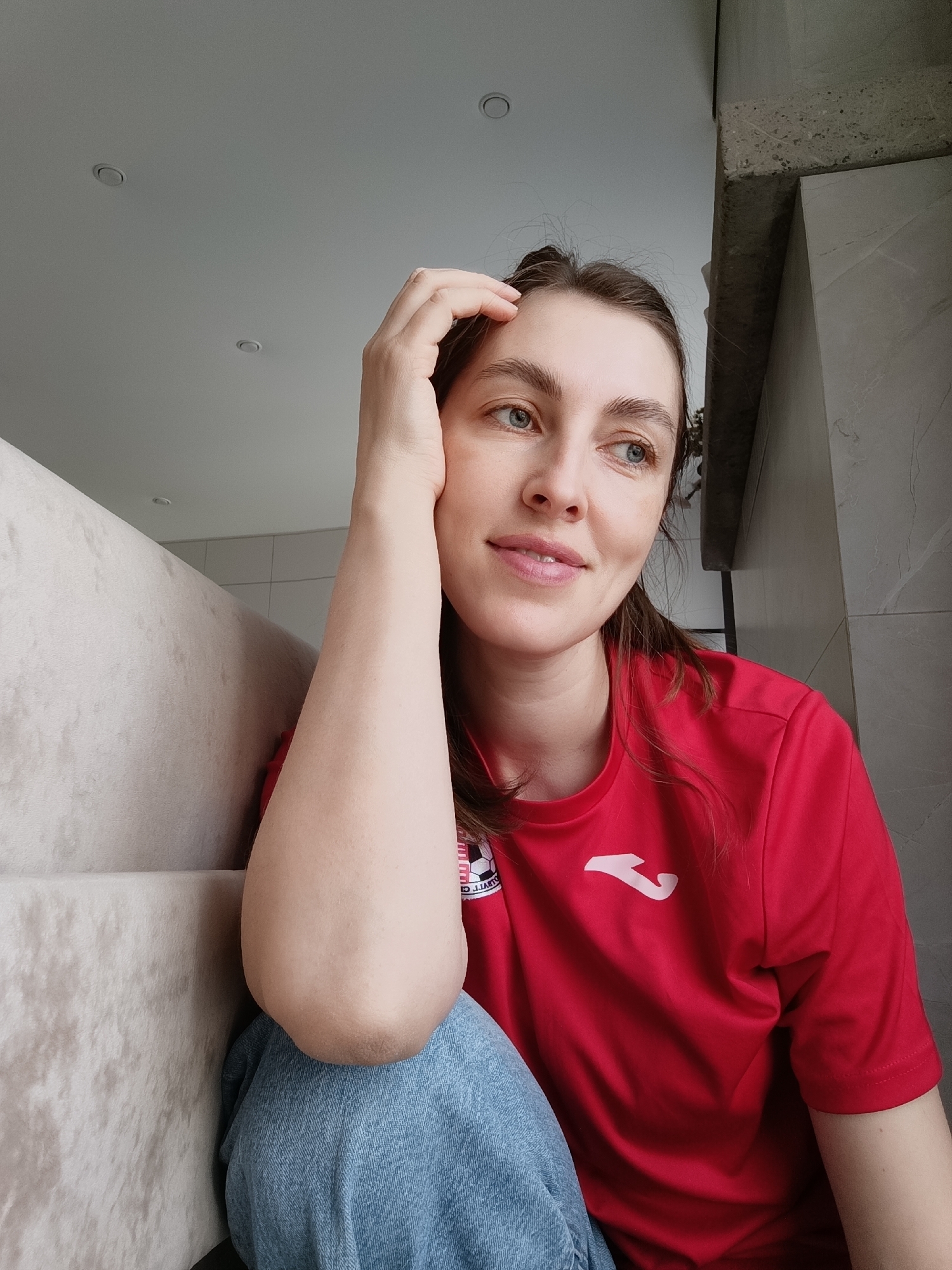 A woman is captured in a close-up, indoor shot, from her head to just below her waist. She has fair skin, light brown hair styled away from her face, and blue eyes, and wears a soft, contemplative smile while gazing upwards and to her right. Her right hand is gently placed on her temple.
She is dressed in a bright red short-sleeved t-shirt, which features a prominent white Joma logo on the chest. To the left of the Joma logo, a circular emblem is partially visible, showing a black and white checkered pattern resembling a soccer ball, with the text "O. JUATT C." discernible along its top curve. She is also wearing light blue denim jeans.
The setting appears to be a modern interior, possibly a living space. To her left, a light-colored, upholstered sofa or cushioned furniture provides a soft backdrop. To her right and behind her, a wall covered in light grey tiles or panels is visible, topped by a raw concrete beam. The ceiling is white and features two recessed light fixtures, indicating a well-lit space, likely during the daytime. The woman's serene expression and relaxed pose contribute to a peaceful and thoughtful atmosphere within this indoor scene in Chaikovsky, Russia.