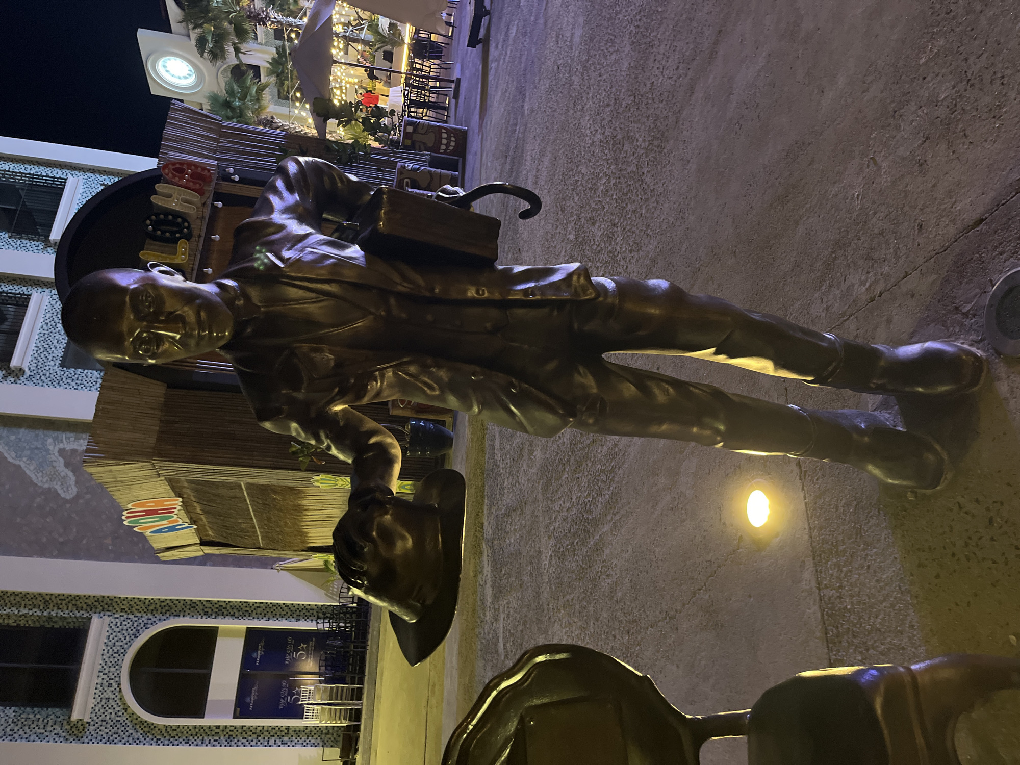A bronze statue of a bespectacled man in a suit, carrying a briefcase and a hat, stands on a paved surface. A building with arched windows and signs that read "PARAGON CASINO" and "THAP CAM DO 5*" is visible in the background, along with outdoor seating and decor. The location is in the city of Bac Nha Trang Ward, Vietnam.