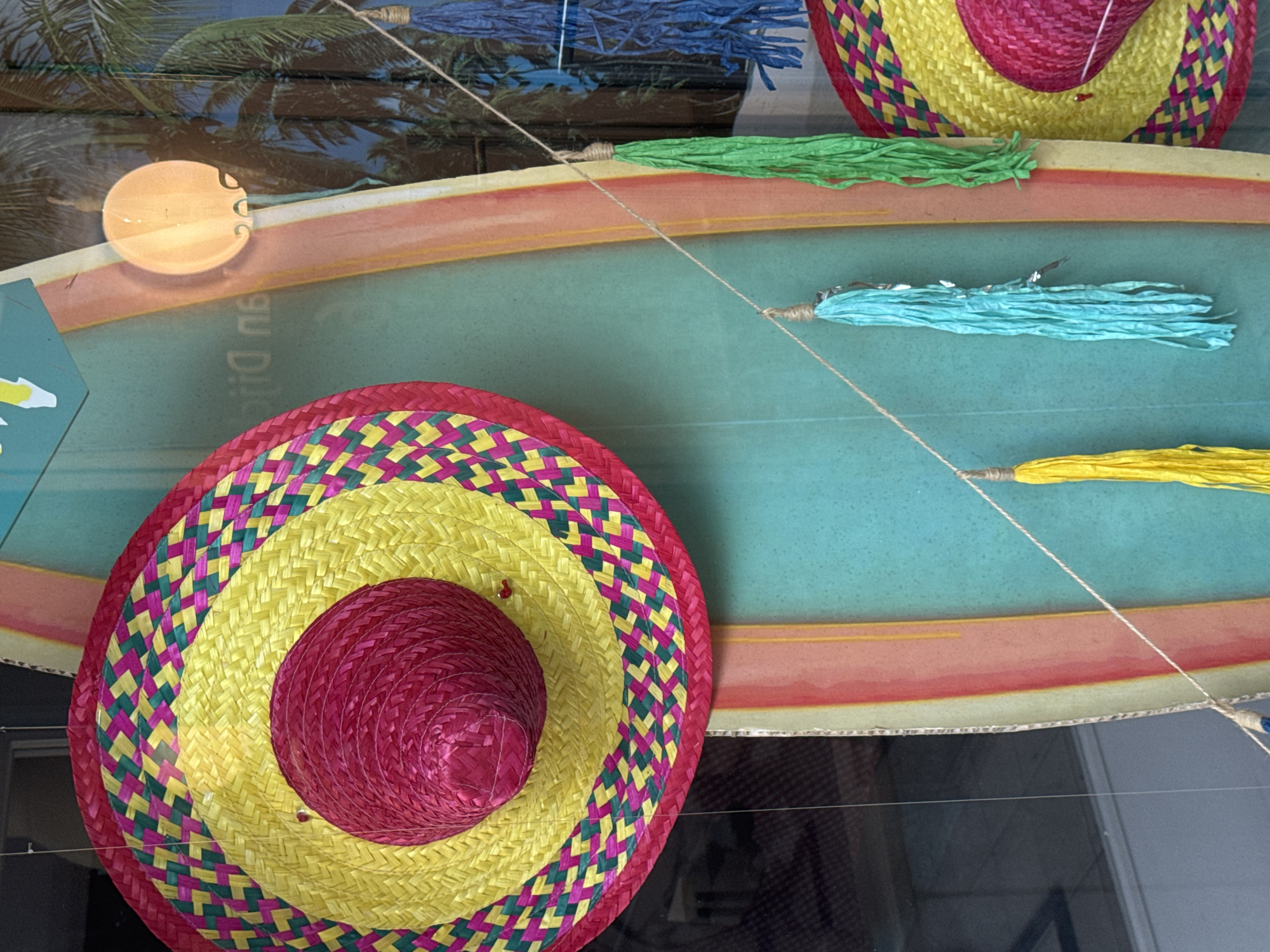 The image shows a close-up of a decorative display, likely in a shop window, featuring Mexican-themed items. Dominating the frame is a large, colorful sombrero with concentric circles of yellow and pink straw, edged with a diamond pattern of pink and green. Below it, part of another similar sombrero is visible on the right. Hanging on thin strings are several bright, textured tassels in shades of yellow, turquoise, and green. In the background, a surfboard-like object with rainbow stripes in pink, orange, yellow, and green stands vertically. Behind the surfboard, a reflection shows palm trees and what appears to be a circular light fixture. The overall impression is festive and summery. The location is a shop window in Strombeek-Bever, Belgium.