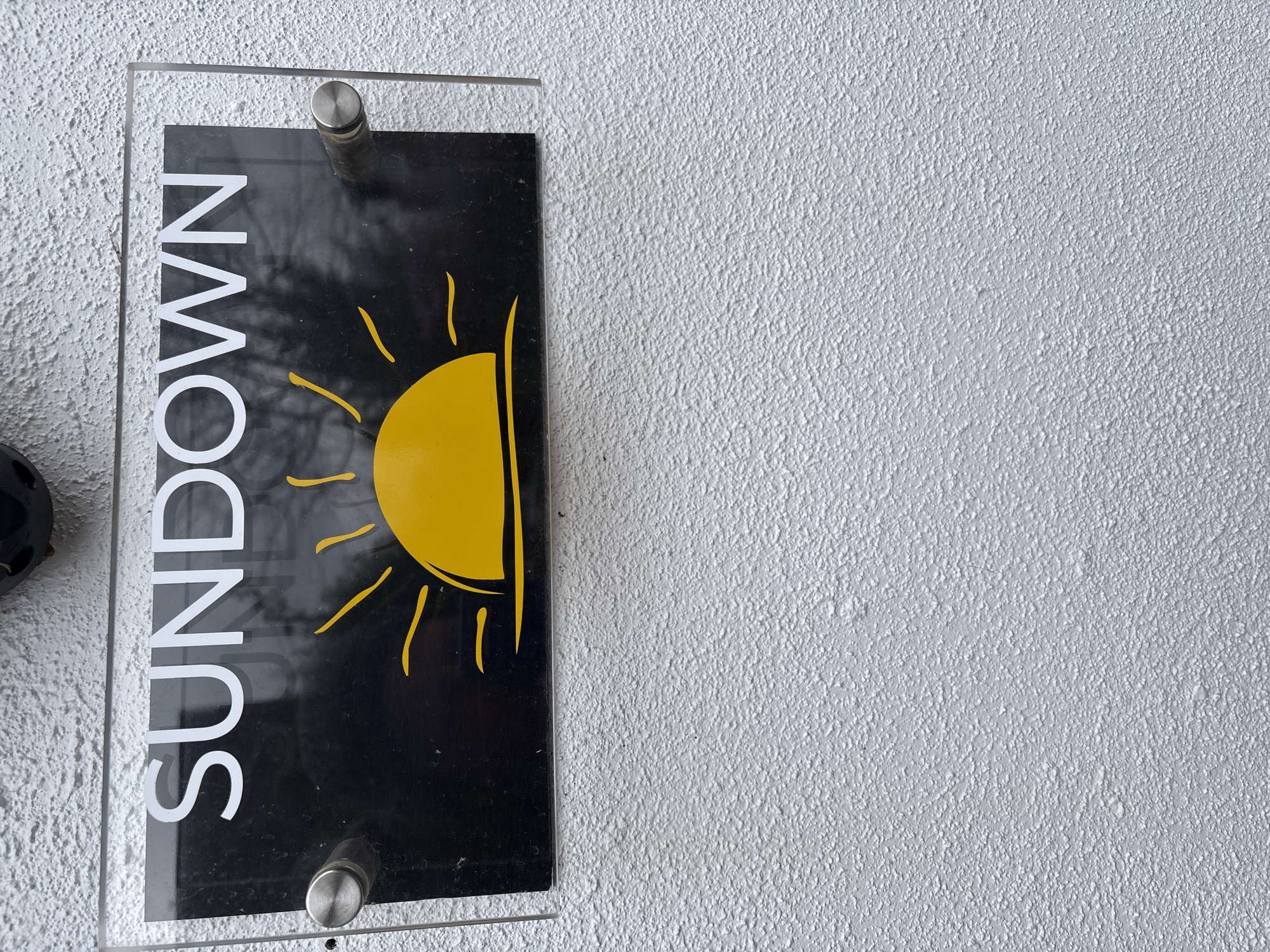 A sign reading "SUNDOWN" with a yellow sun graphic is mounted on a white, textured wall. The sign is black with white lettering, enclosed in a clear acrylic frame affixed to the wall with metal standoffs. Part of an unidentifiable black object is visible on the left edge of the frame. The background of the sign includes reflections of trees. The photograph was taken in Longden, United Kingdom.