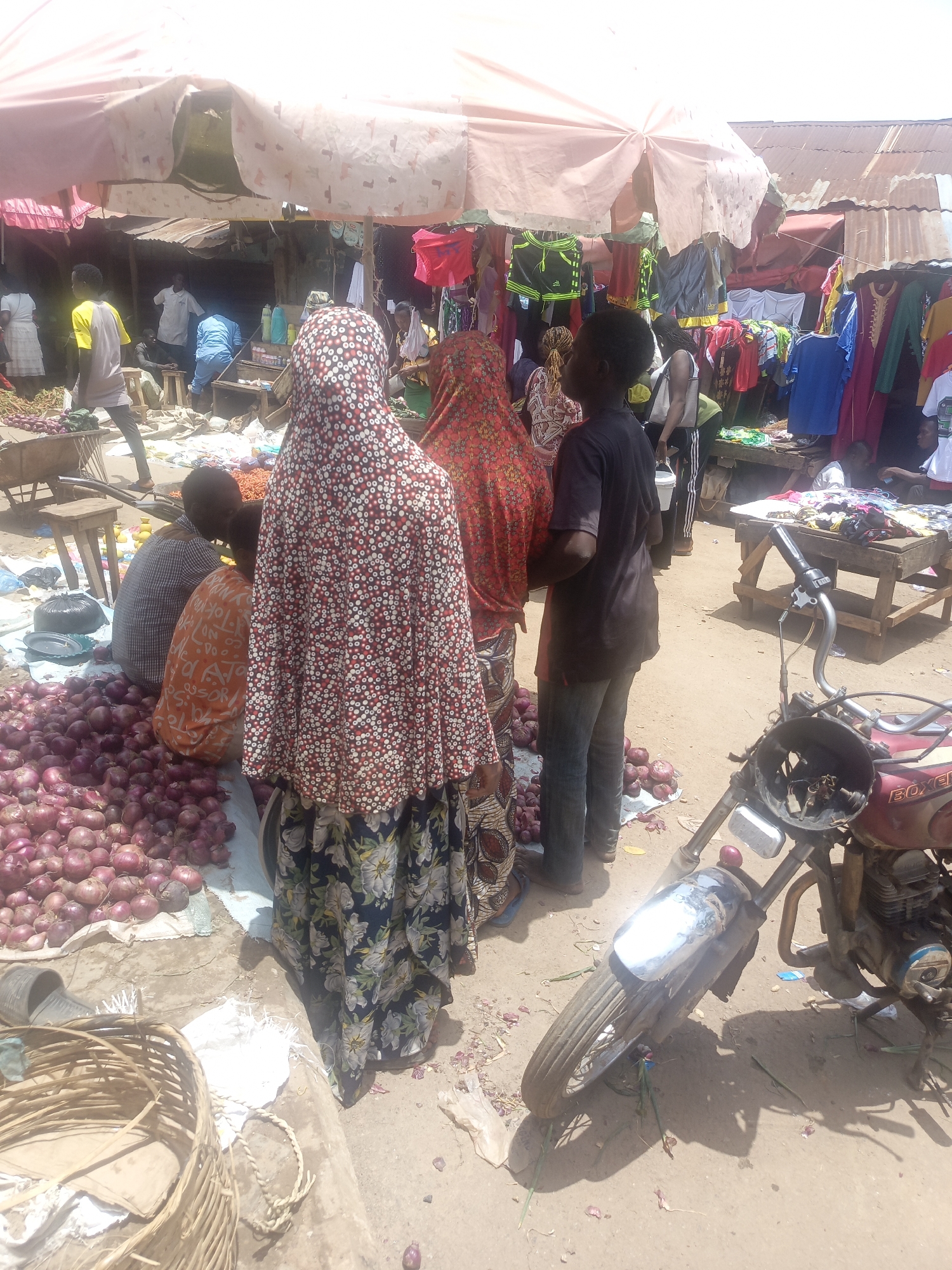The image shows a bustling market scene in an unknown city. Several people are gathered around a large pile of red onions, presumably for sale. The buyers are mostly women, some wearing colorful headscarves and long dresses. A young man in a dark shirt stands among them. In the background, there are stalls selling clothes and other merchandise. A motorcycle is parked on the side, partially obstructing the view. The ground is dusty, and there is an umbrella providing shade.