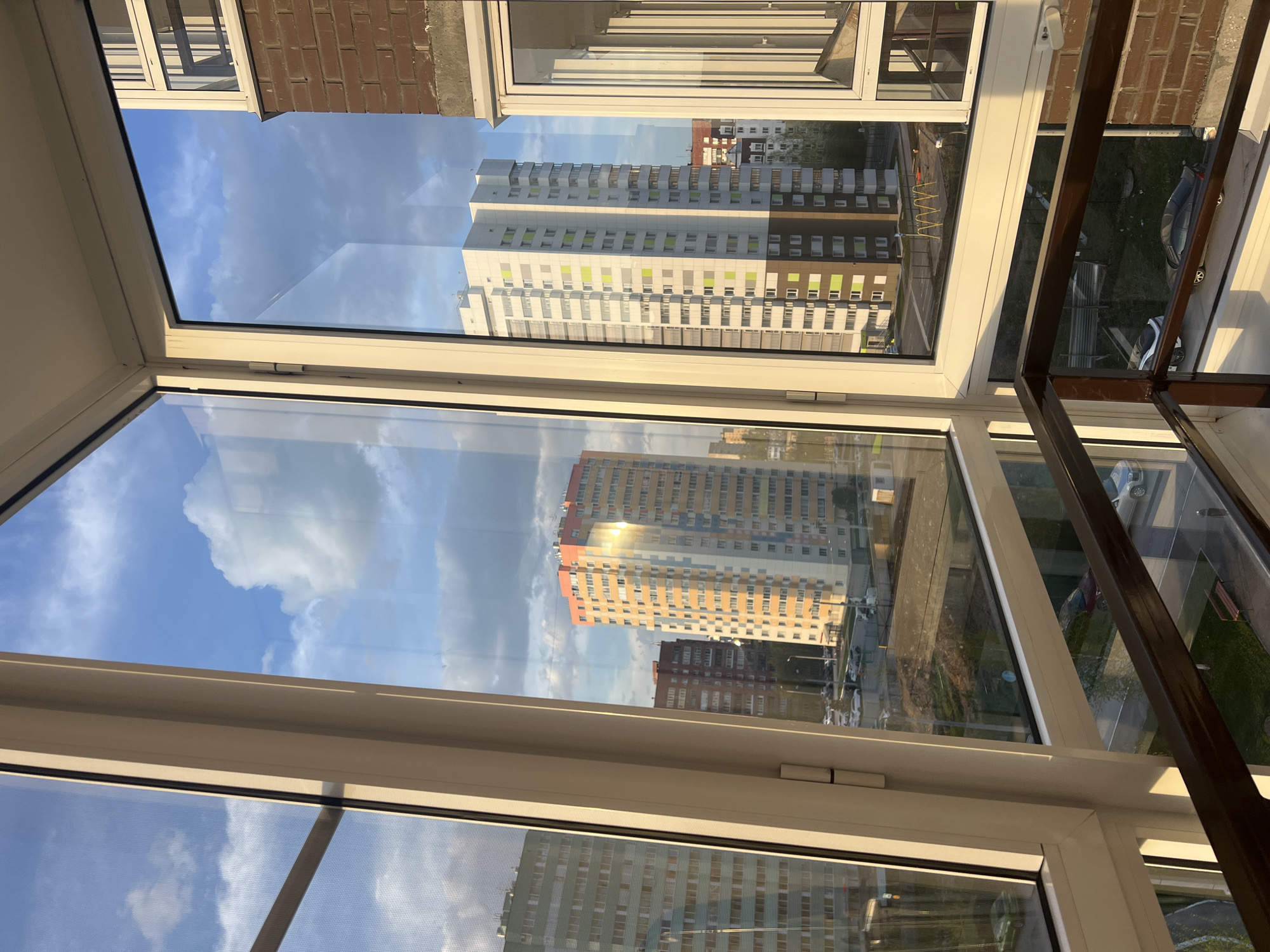 The image, taken through a window, offers a view of an urban landscape with buildings and sky. The window frame itself is white and multi-paned. Outside, various high-rise apartment buildings are visible in the background. One building is predominantly white with green accents, while another has a reddish-orange hue. The sky features a combination of blue and white clouds. Part of a metal railing is visible on the right side. The scene appears to be in Perm, Russia.