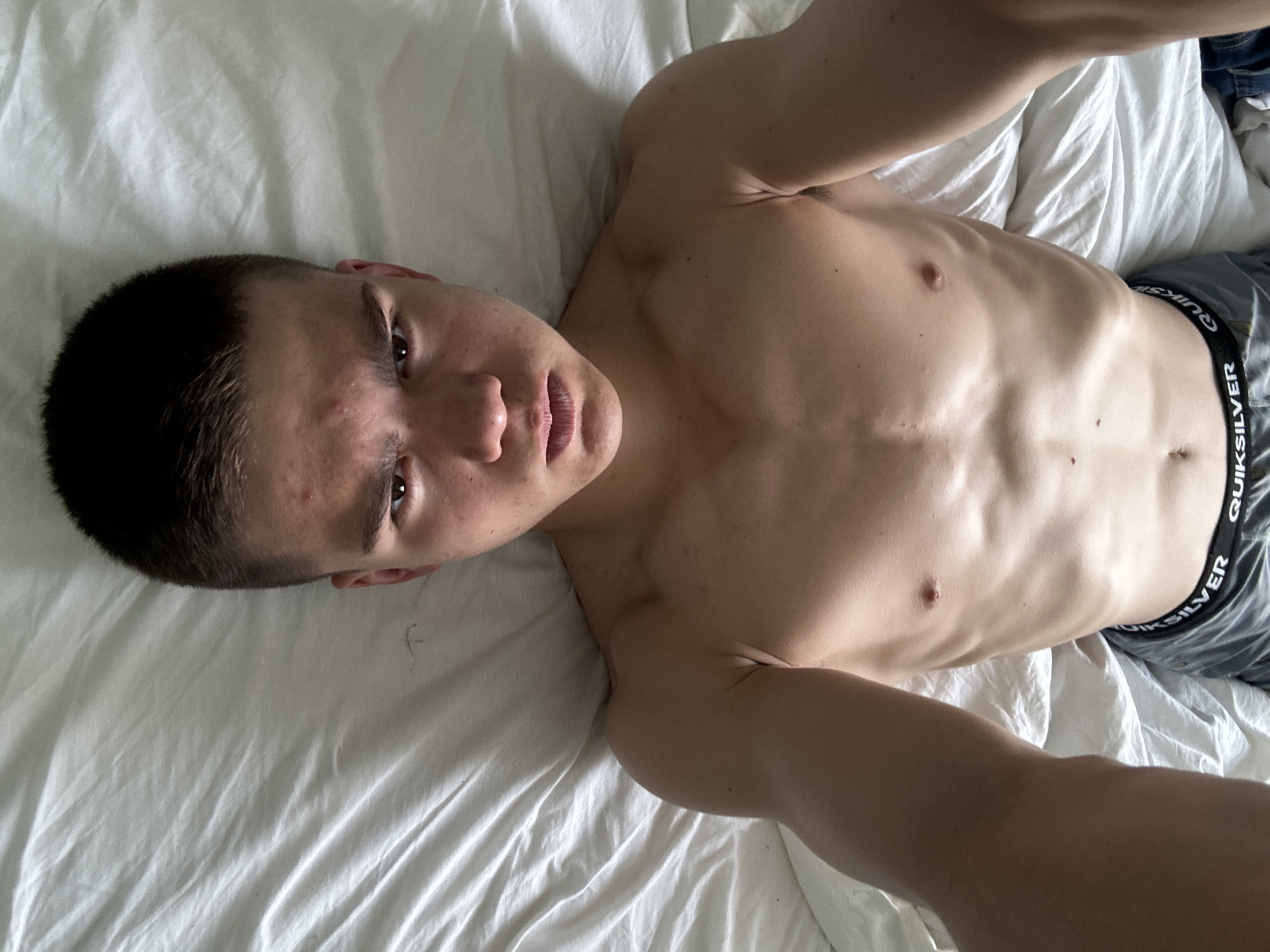 A young man is lying on a white bed. He is shirtless, and wearing gray Quiksilver underwear and jeans. The background consists of a white sheet.