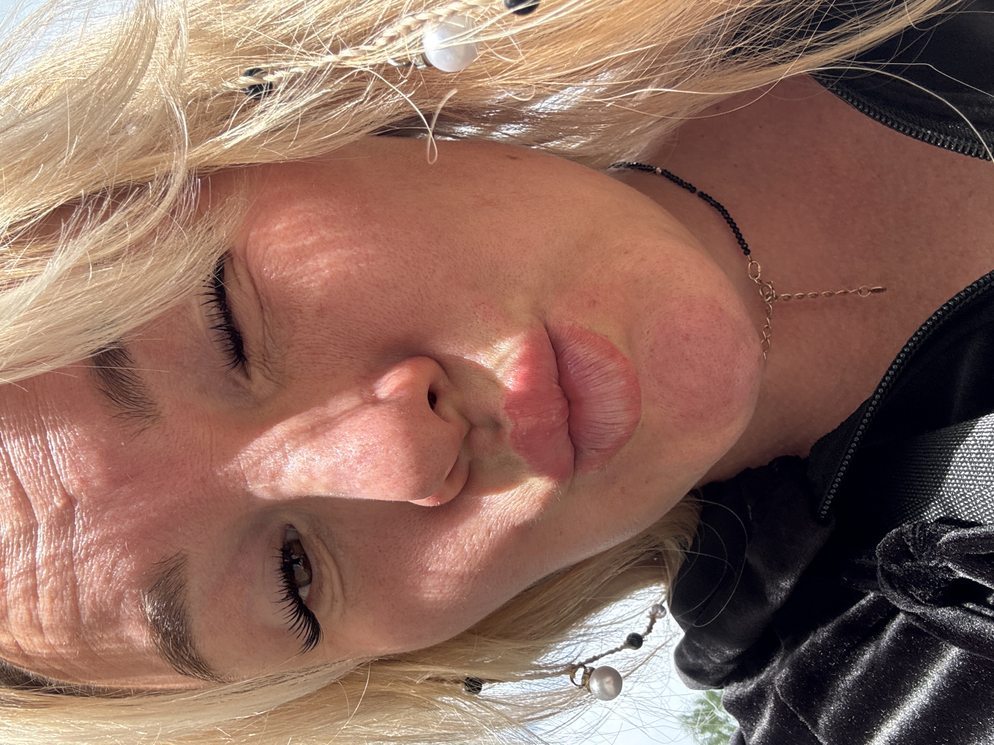 The image shows a close-up selfie of a young woman with blonde hair and light skin. She is winking and puckering her lips, and her hair is partially covering her face. She is wearing a black velvet top and a black beaded necklace. She has large false eyelashes. The background is blurred and appears to be outdoors, with some greenery and a bright sky. The lighting suggests it is daytime. No specific details about Chastyye, Russia, are visible in the image.