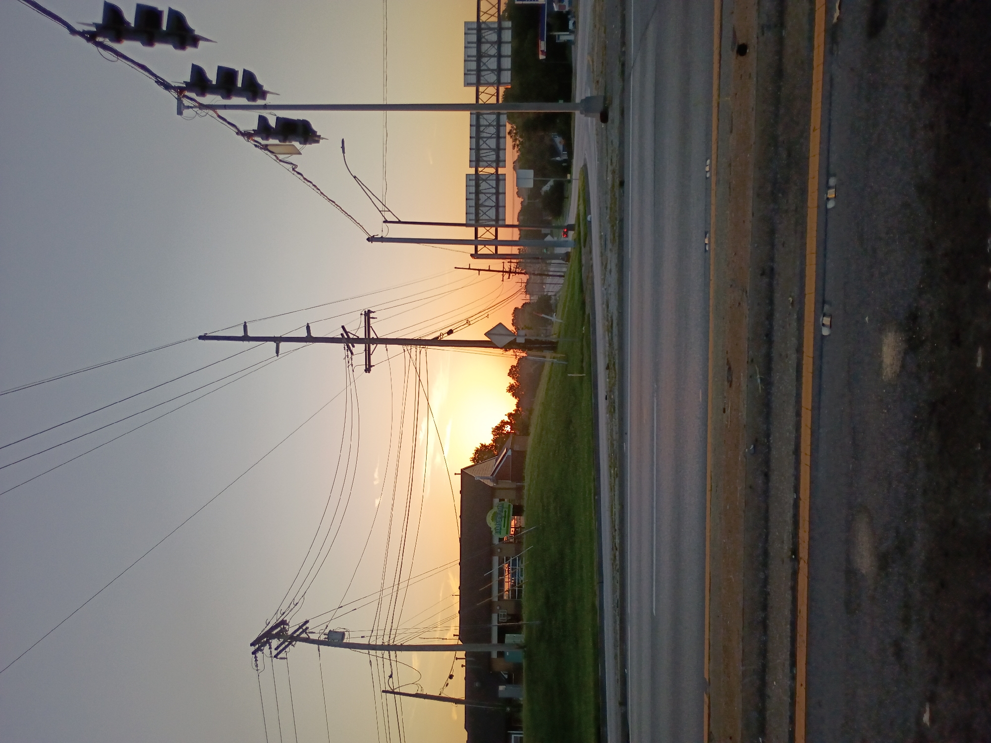 This media file captures a sunset scene in Frankfort, United States of America. The image is oriented vertically, with the top of the frame showing a clear sky transitioning to warm hues of orange and yellow as the sun sets. Several traffic lights are visible in the upper left, silhouetted against the bright sky. Numerous telephone poles and power lines crisscross the frame, indicating an urban or suburban setting.
In the mid-ground, a building with a sign that reads "WALK-IN DENTAL" is partially visible, surrounded by a grassy area. The road, with visible yellow lane markings, runs along the right side of the frame, appearing wet, suggesting recent rain or dew. The overall mood is calm and serene, enhanced by the soft lighting of the sunset. The time of day is clearly evening, and the weather appears clear, though the wet road implies some moisture in the air. The presence of traffic lights and the road suggests a public thoroughfare.