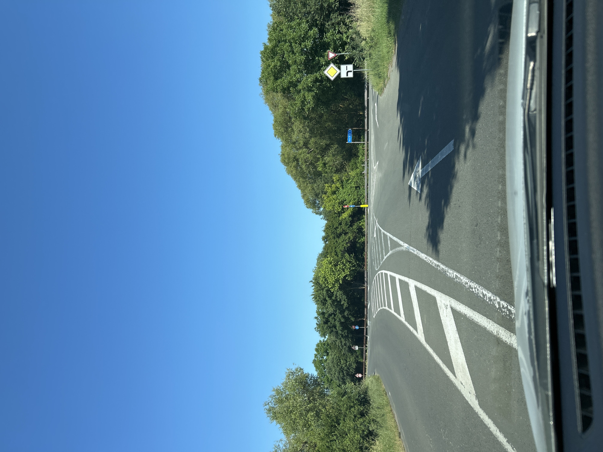 The image shows a clear, sunny day with a bright blue sky. A road diverges into two paths, with white lane markings indicating the directions. Lush green trees and vegetation line the sides of the road. Several road signs are visible, including yield signs and directional signs. The perspective is from inside a vehicle, looking out through the windshield. The scene depicts a typical road intersection in a rural or semi-rural area. No people are visible in the image.